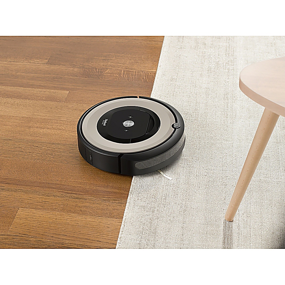 Un aspirapolvere Roomba su un pavimento in legno e un tappeto. Un tavolino è sulla destra. Il Roomba è nero e argento.