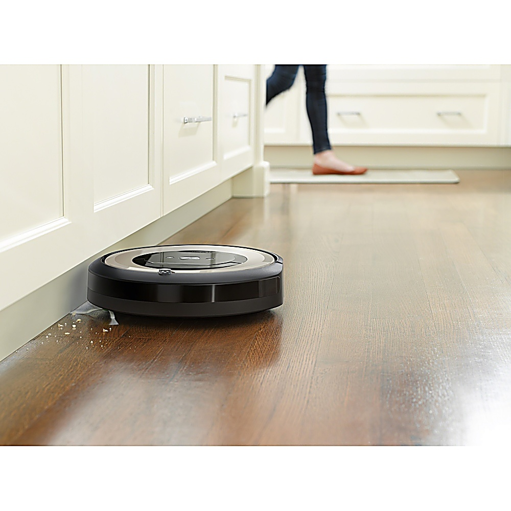 Un aspirapolvere robot nero su un pavimento di legno. Una persona cammina sullo sfondo. Ci sono armadi bianchi a sinistra.