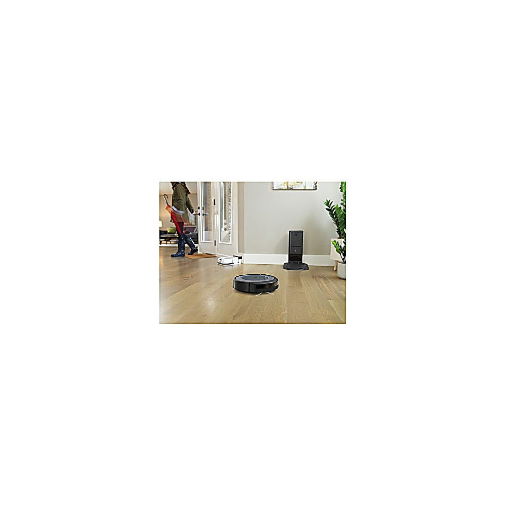 Ein Roboter-Staubsauger auf Holzboden. Eine Person geht durch eine Tür. Eine Ladestation und eine Pflanze sind im Hintergrund.