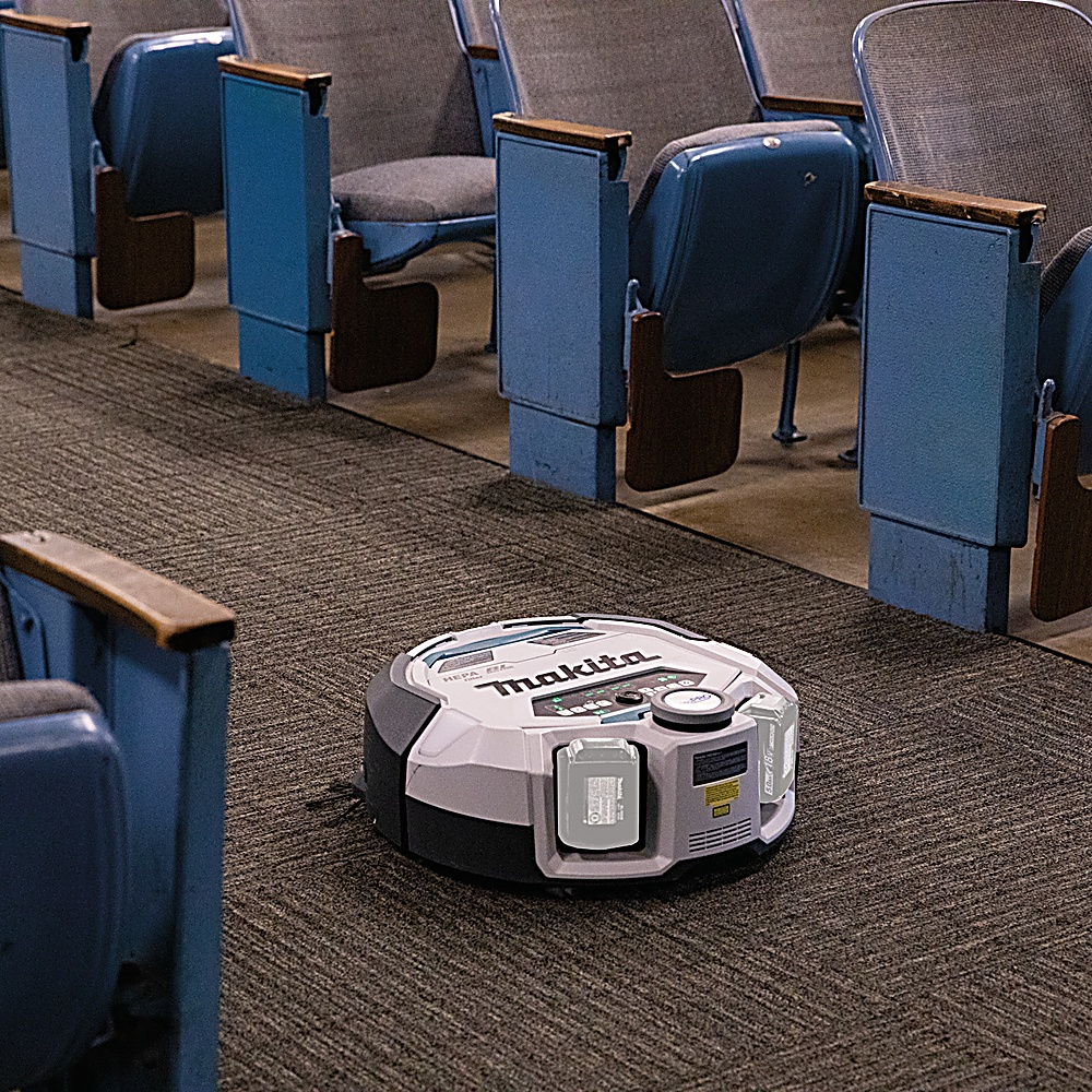 Un aspirapolvere robot Makita su un tappeto in un auditorium. Sedili blu con braccioli in legno sullo sfondo.