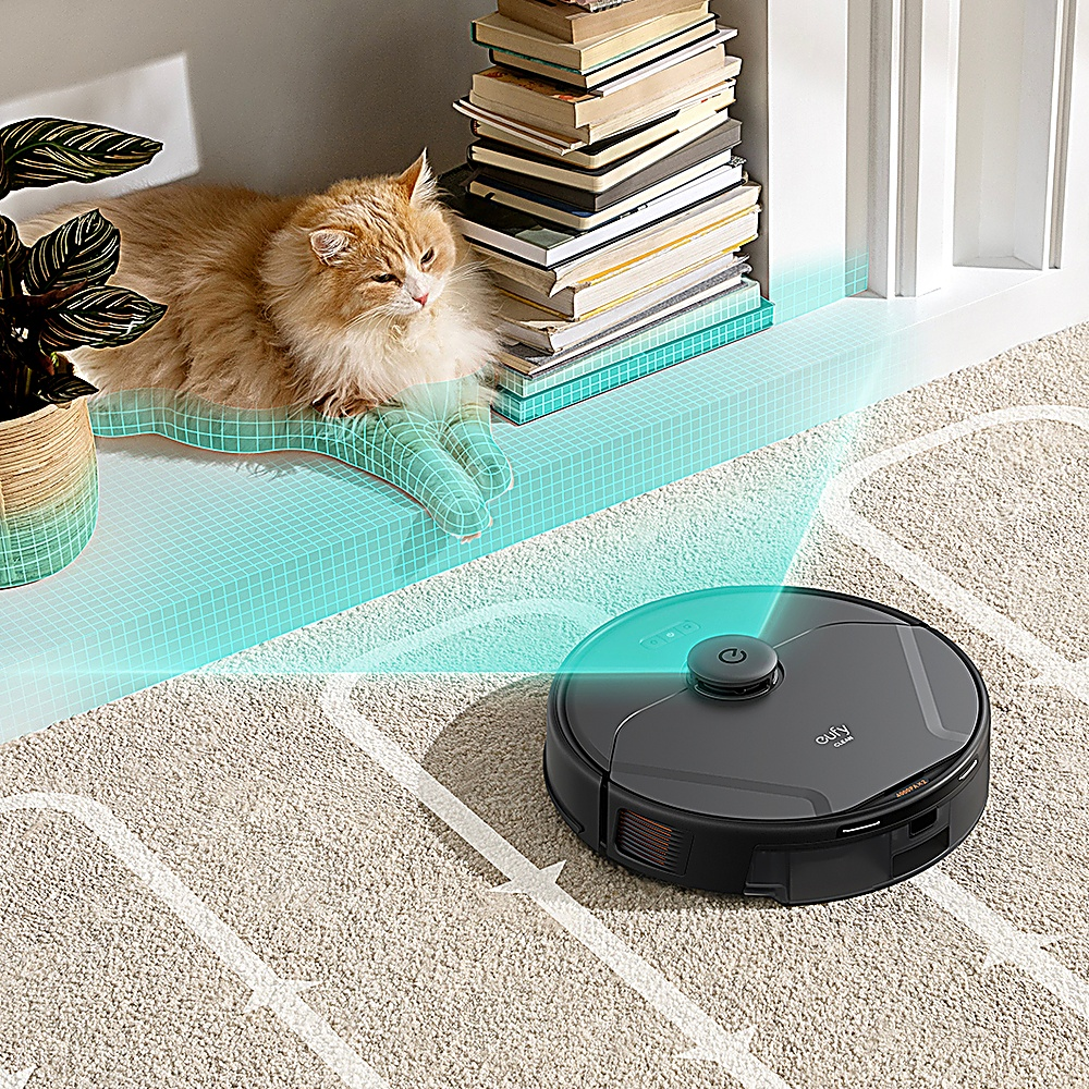 Robot sprzątający na dywanie, kot leży na platformie w pobliżu książek i rośliny.