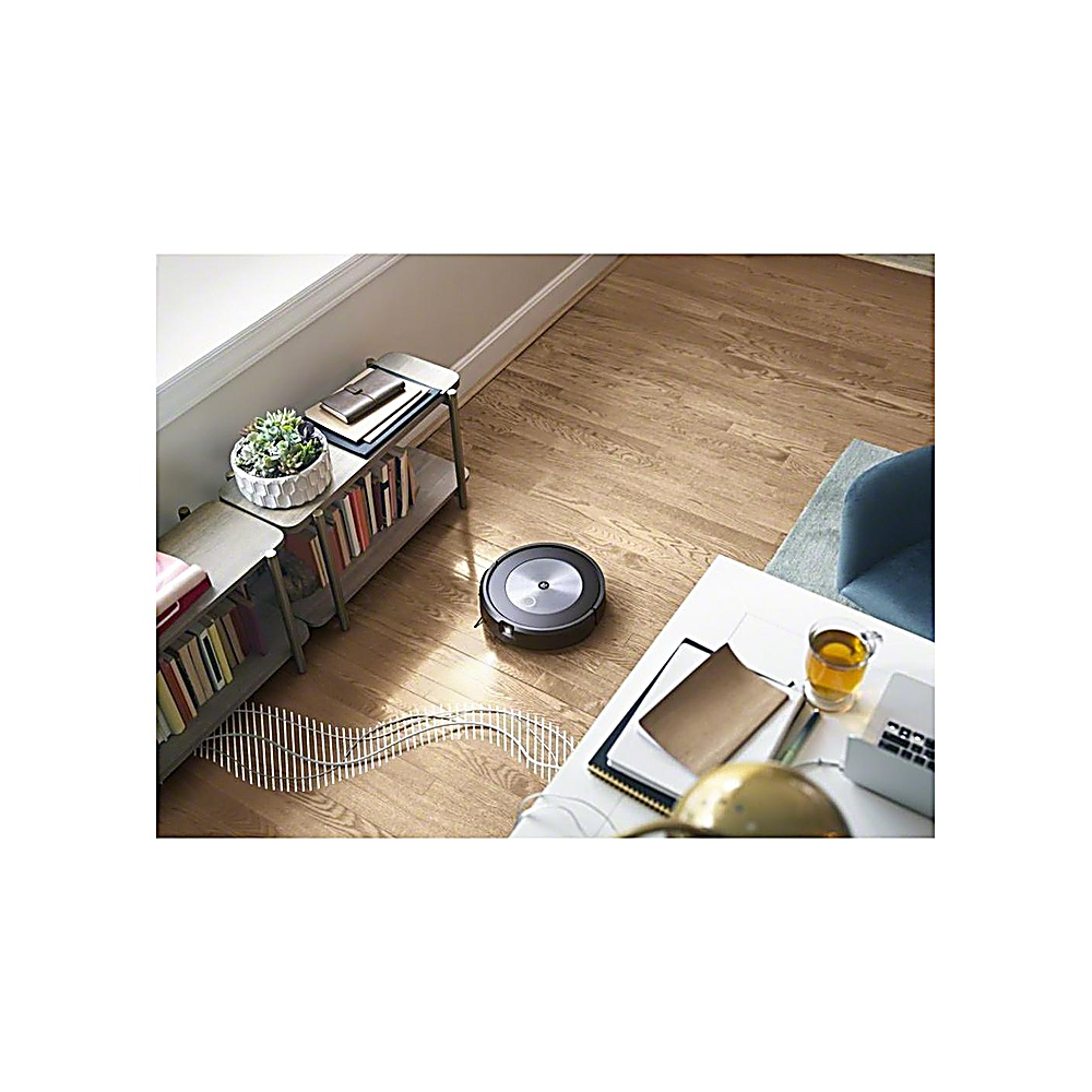 Un robot aspirapolvere pulisce un pavimento in legno. C'è un tavolo con un computer portatile e tè. Sono visibili librerie e un tavolino con piante.