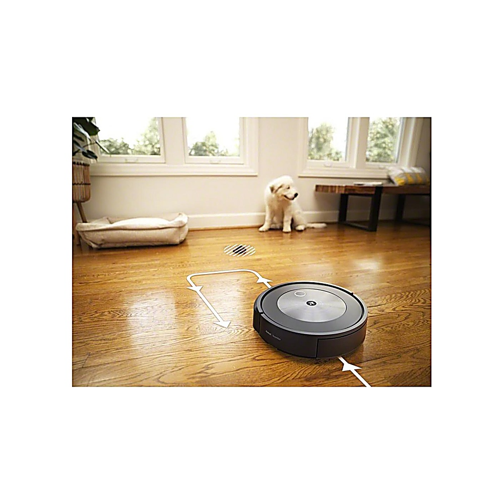 Un robot aspirapolvere su un pavimento in legno. Un cane è seduto sullo sfondo vicino a una finestra. È visibile anche una cuccia.