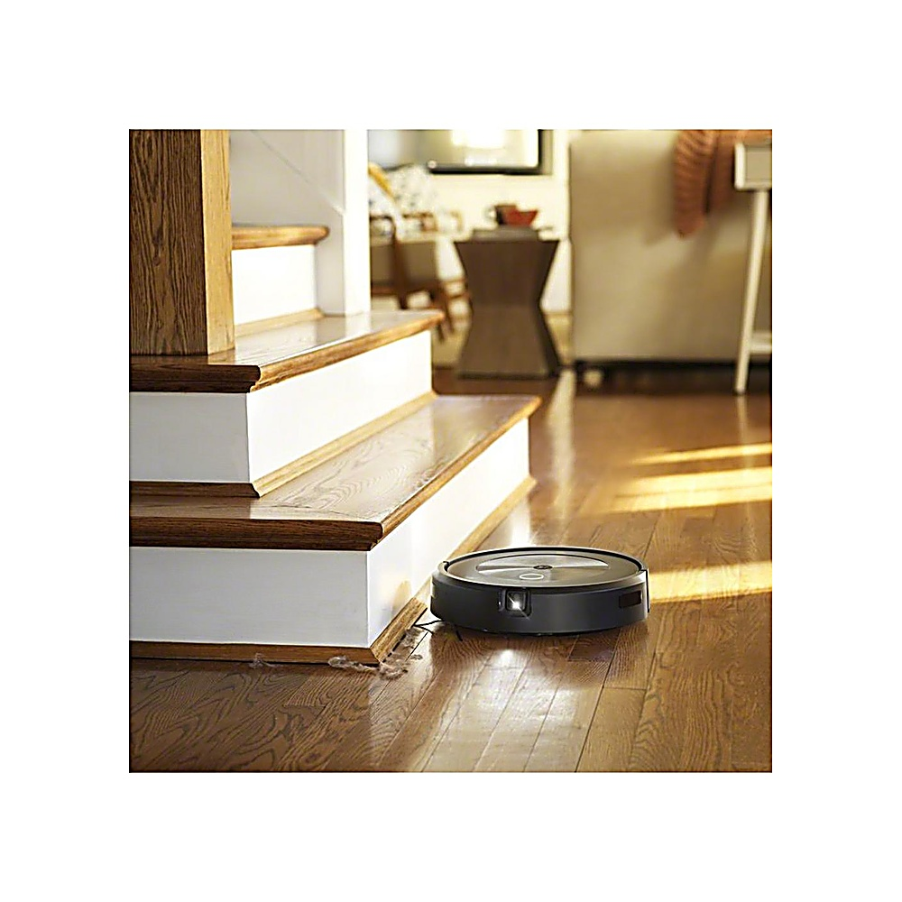 Un aspirapolvere robot nero su un pavimento di legno vicino a una scala. Le scale hanno alzatine bianche e gradini marroni.