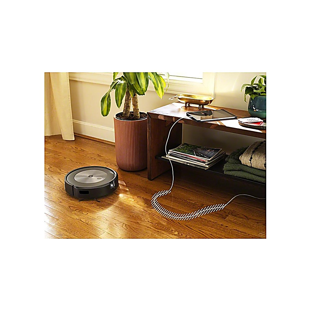 Un robot aspirapolvere su un pavimento in legno. Una pianta, un tavolo di legno con riviste e una finestra sullo sfondo.