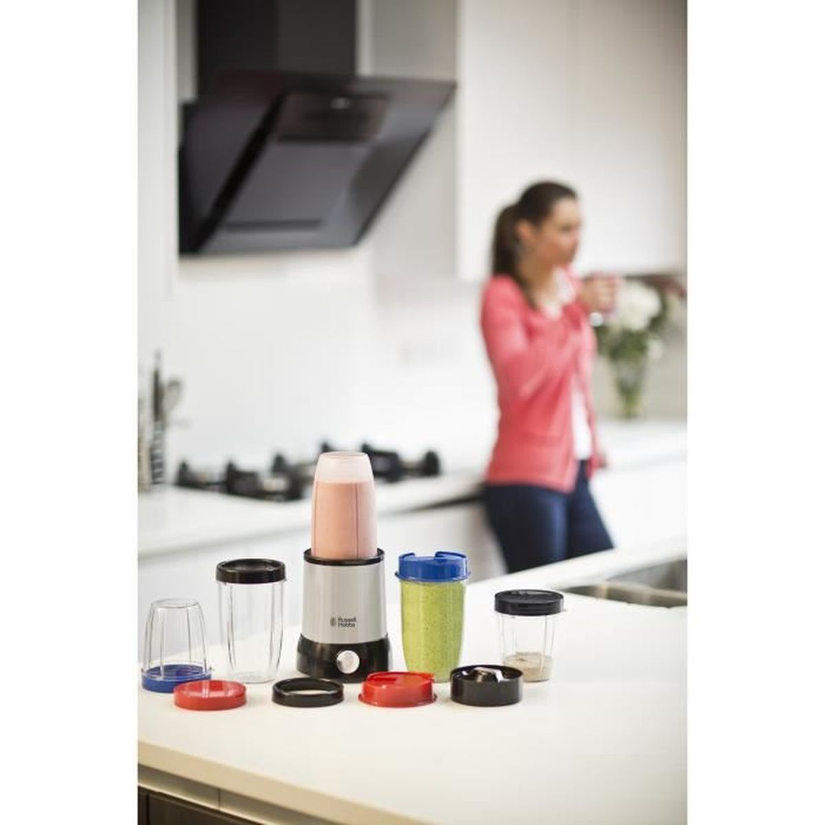 Bir mutfak sahnesi, bir blender ve çeşitli kaplar. Arka planda bir kadın var. Blender beyaz bir tezgahta duruyor.