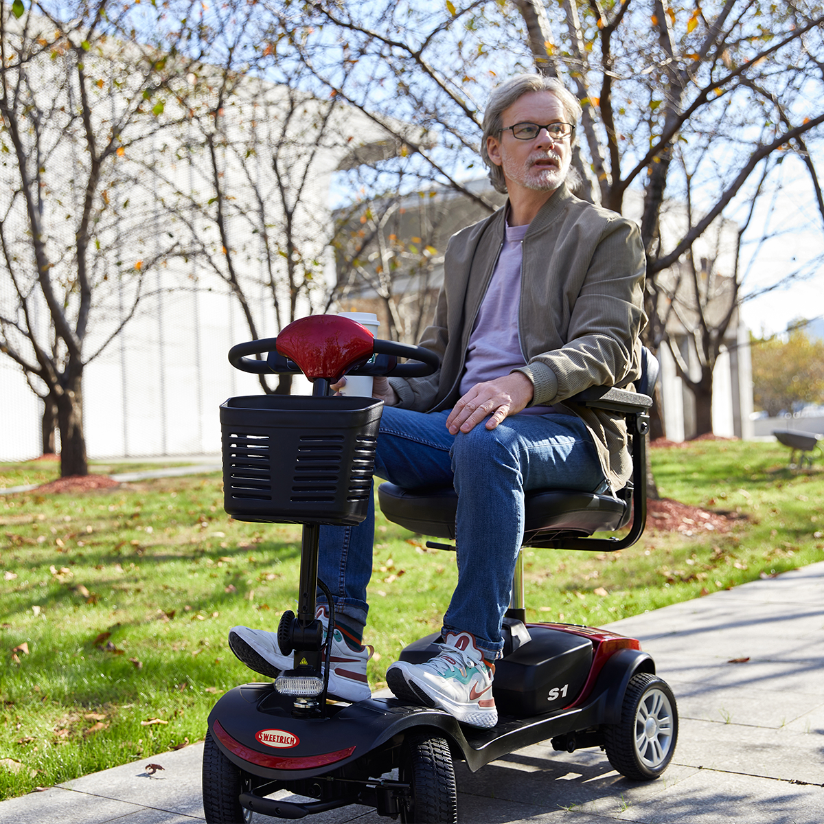 Mężczyzna na skuterze inwalidzkim na zewnątrz. Nosi okulary, kurtkę i dżinsy. W tle drzewa i trawa.