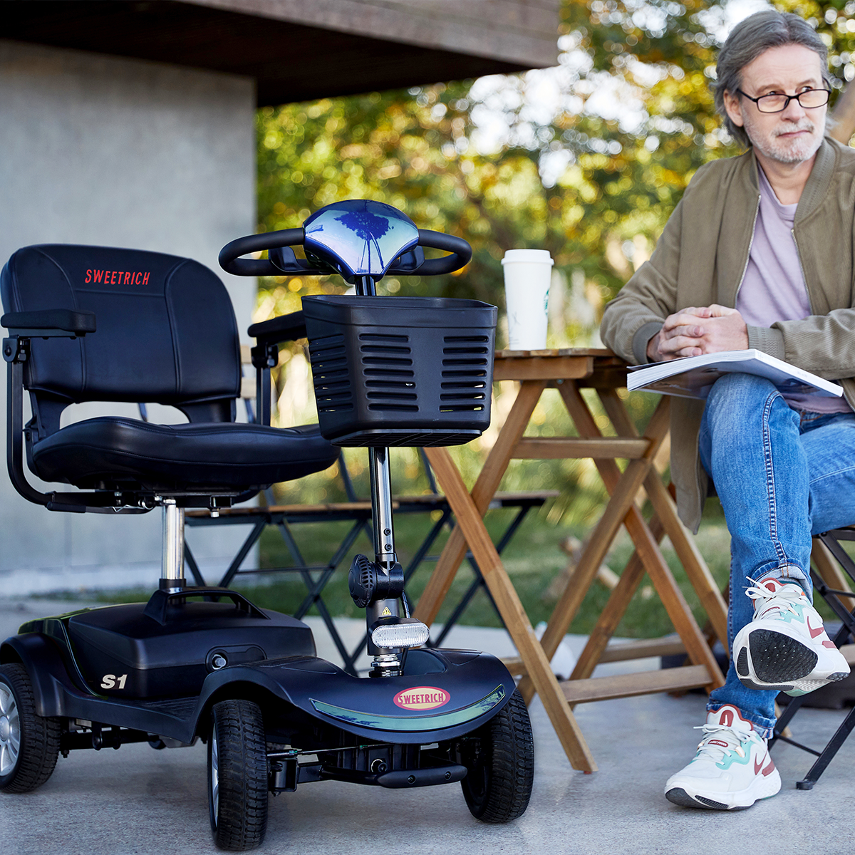 Mężczyzna siedzi obok czarnego skutera. Obok niego stół i filiżanka kawy. Ma okulary i dżinsy.
