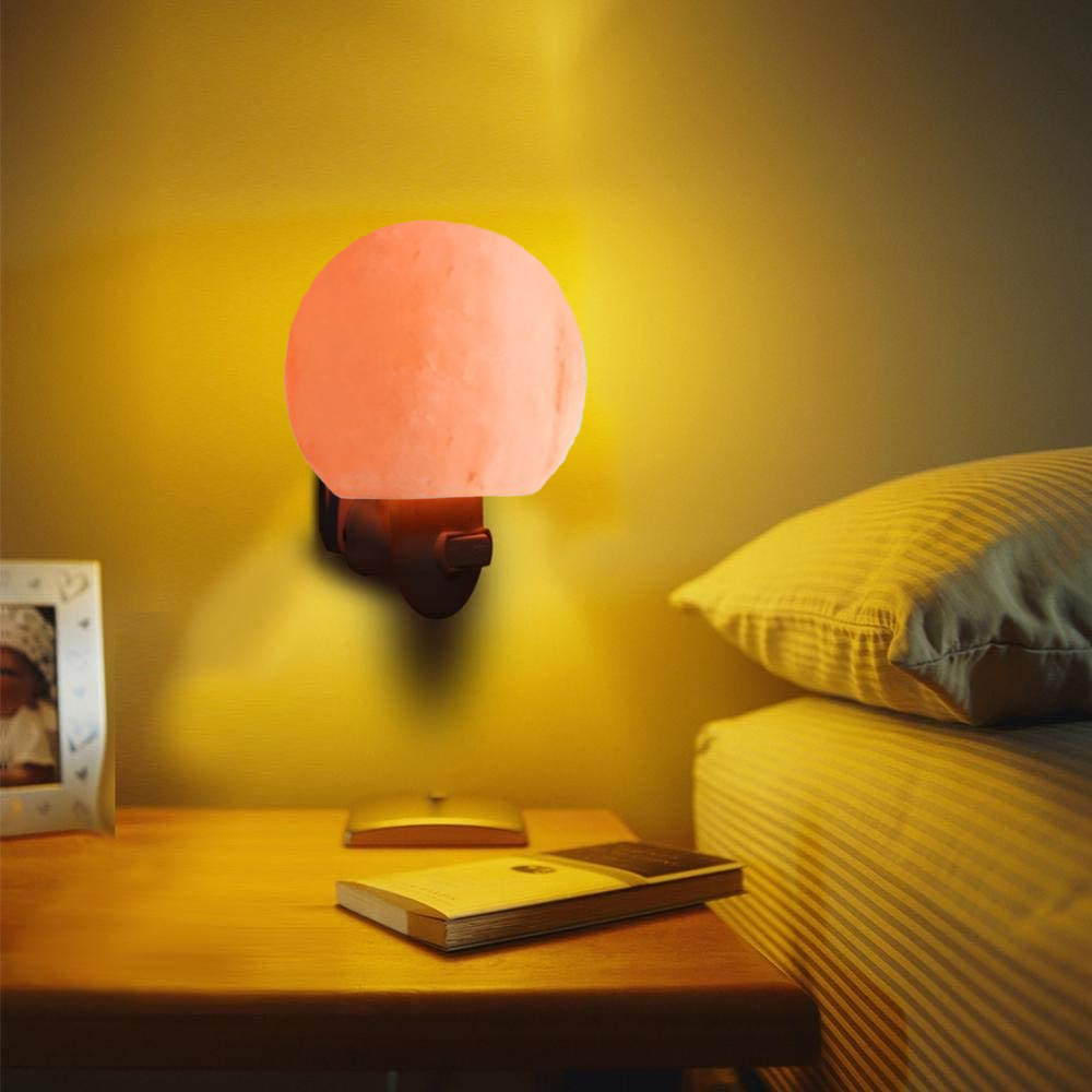 Eine Salzkristalllampe beleuchtet ein Schlafzimmer. Auf einem Nachttisch liegen ein Buch und ein Fotorahmen. Im Hintergrund ein Bett mit Kissen.