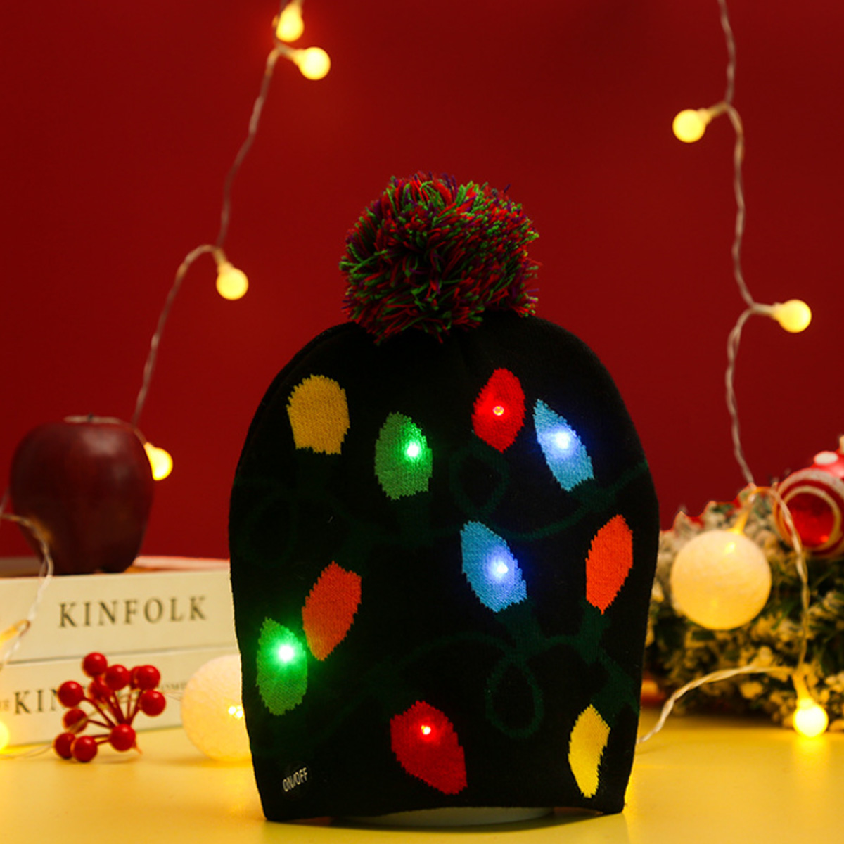 Eine schwarze Mütze mit bunten Weihnachtslichtern. Sie hat einen Bommel und steht auf einem Tisch mit anderen Dekorationen und Lichtern im Hintergrund.