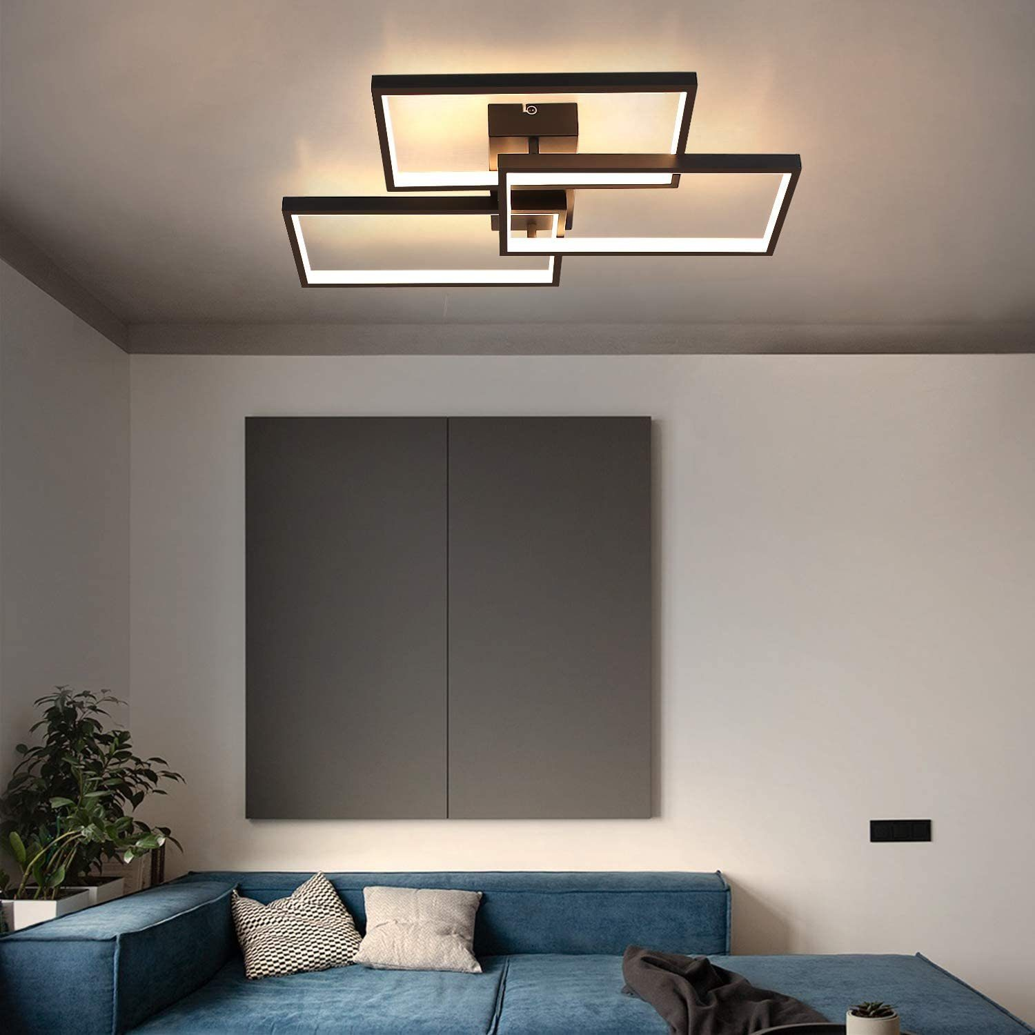 Moderno lampadario a soffitto con forme rettangolari. Sotto, un divano blu con cuscini, una pianta e un muro grigio.