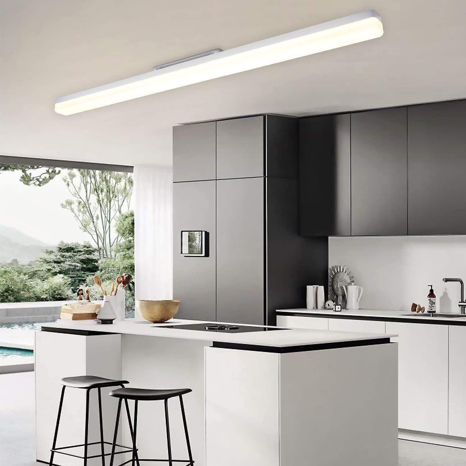 Cucina moderna con isola bianca, sgabelli neri, armadi grigi. Un lungo apparecchio illumina lo spazio. Vista dalla finestra.