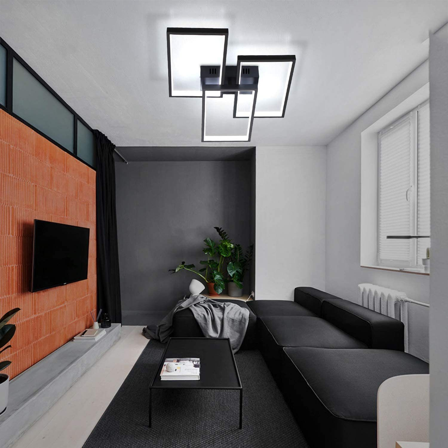 Soggiorno con divano nero, tavolo, muro di mattoni, TV e luci a soffitto. Sono visibili anche piante e una finestra.