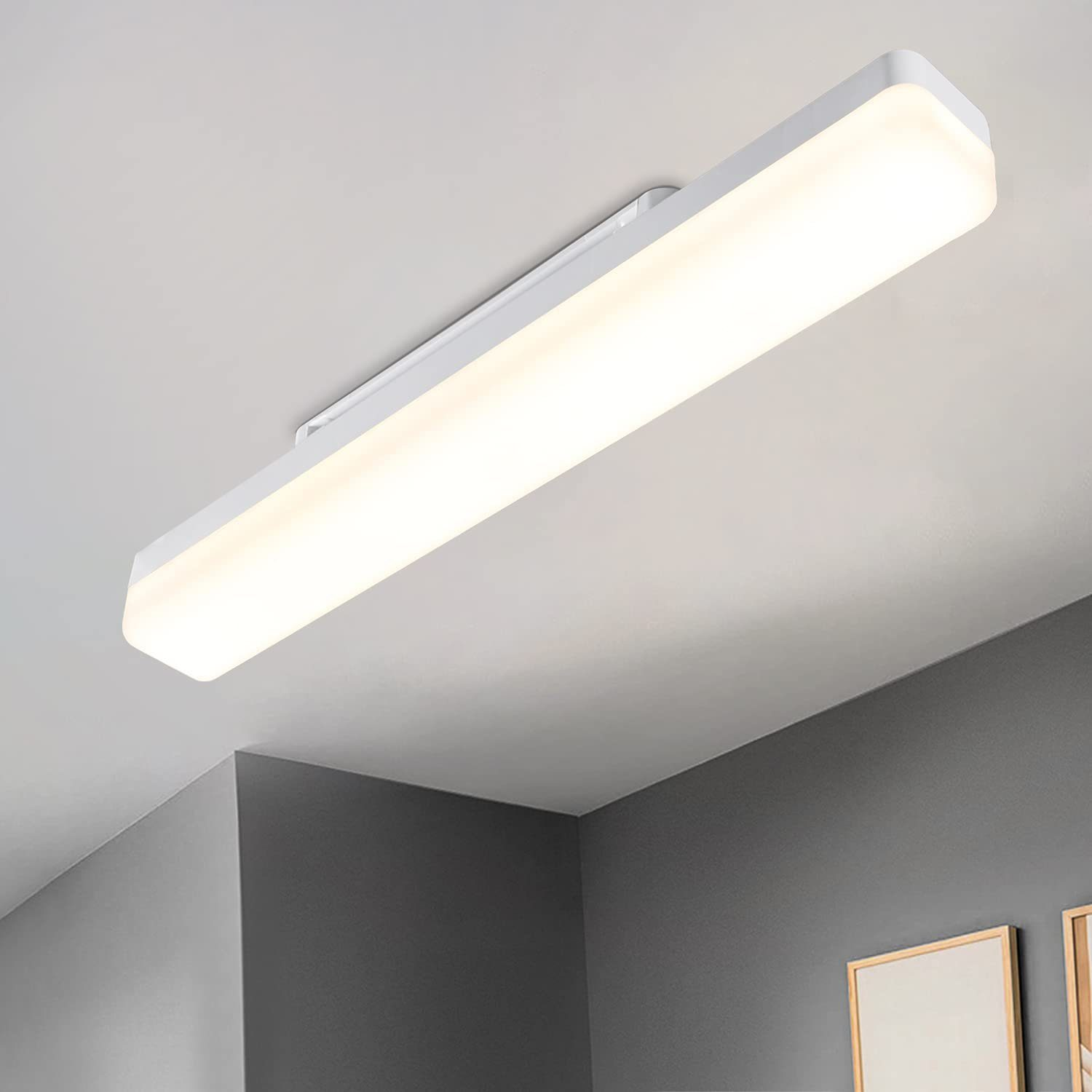 Biała prostokątna lampa sufitowa jest zainstalowana na białym suficie. Widoczne są szare ściany. Obraz w ramie wisi na ścianie.
