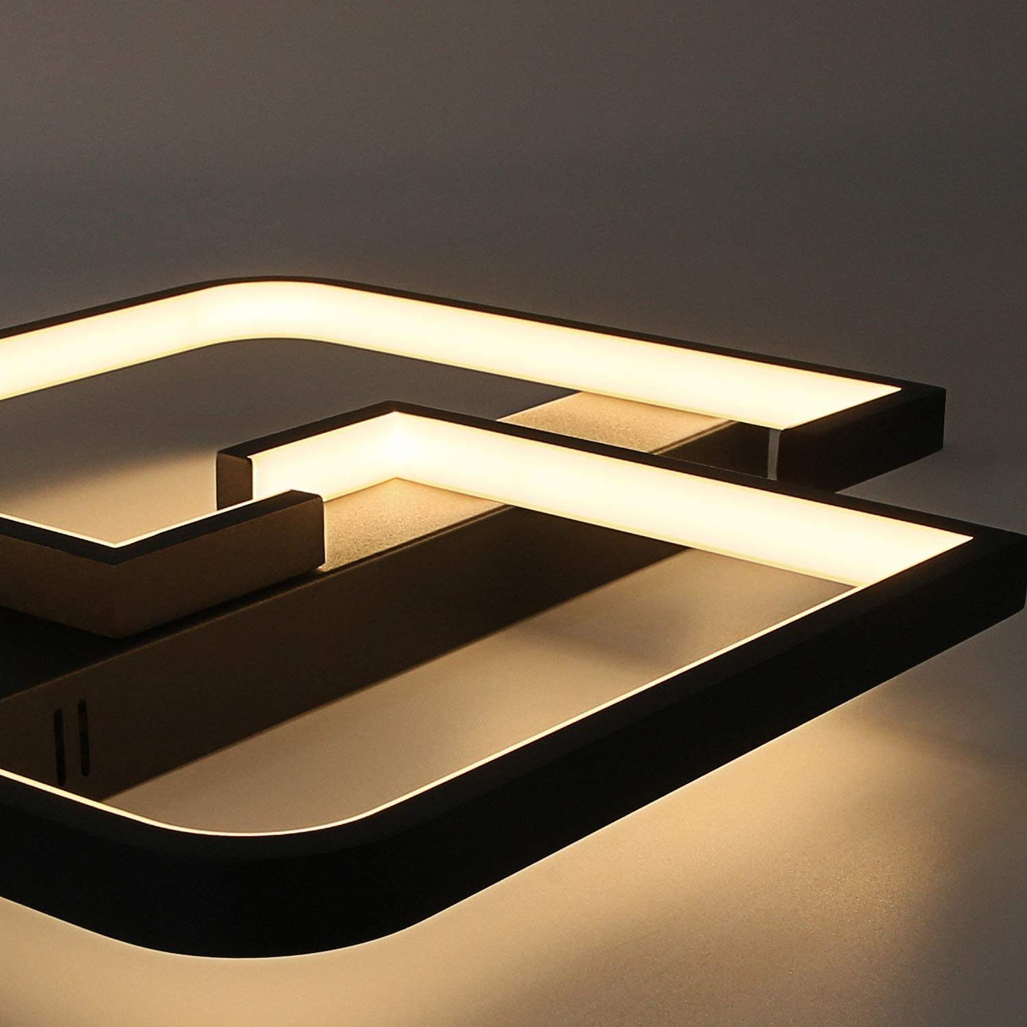 Lampada da soffitto geometrica moderna con cornice nera e luce bianca calda. Ha una forma quadrata ed è montata su una parete grigia.