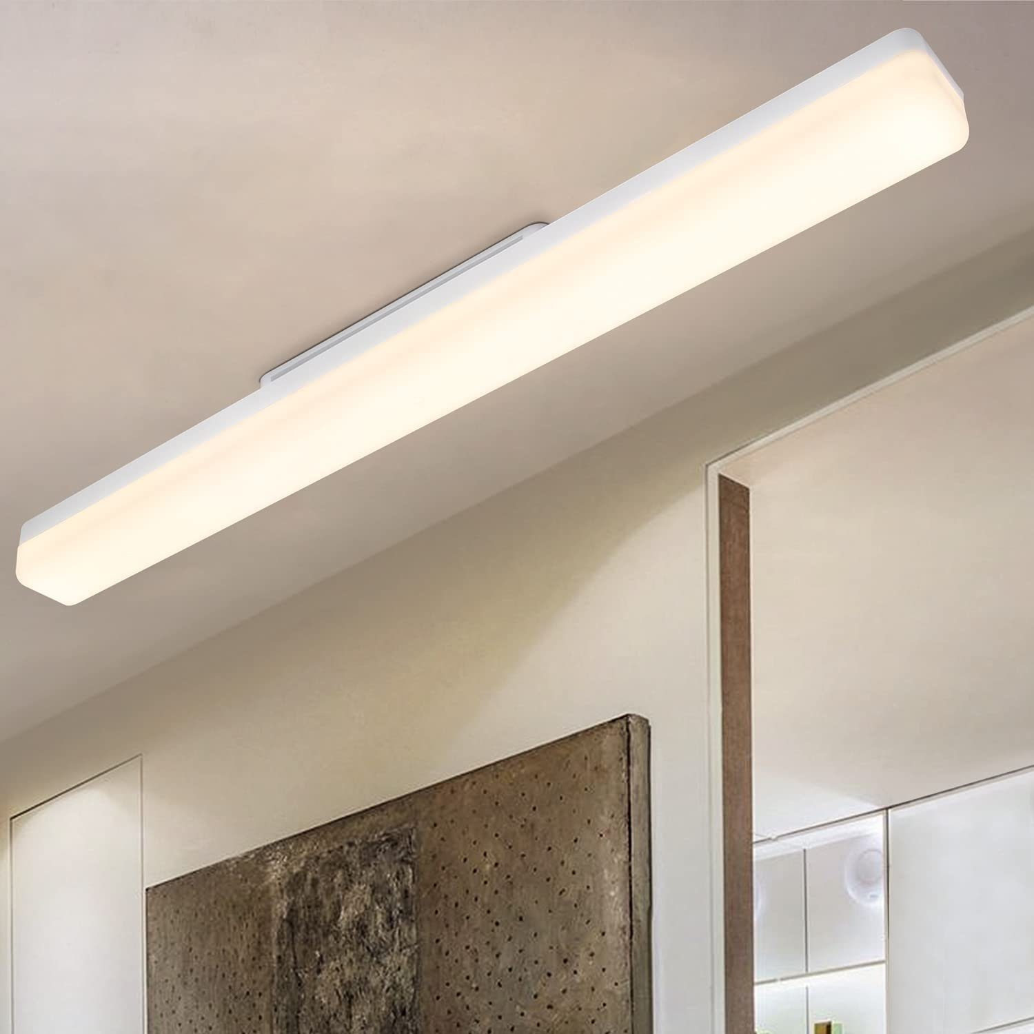 Una lunga lampada da soffitto rettangolare bianca illumina una stanza. Sotto, un'opera d'arte strutturata è appesa al muro, vicino a una porta.