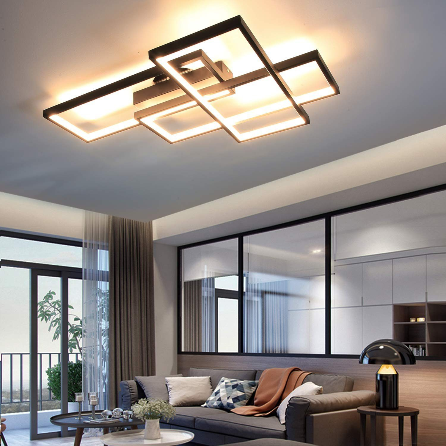 Un soggiorno moderno con una lampada da soffitto geometrica. Divano grigio, finestre e tavolino con lampada sono visibili.