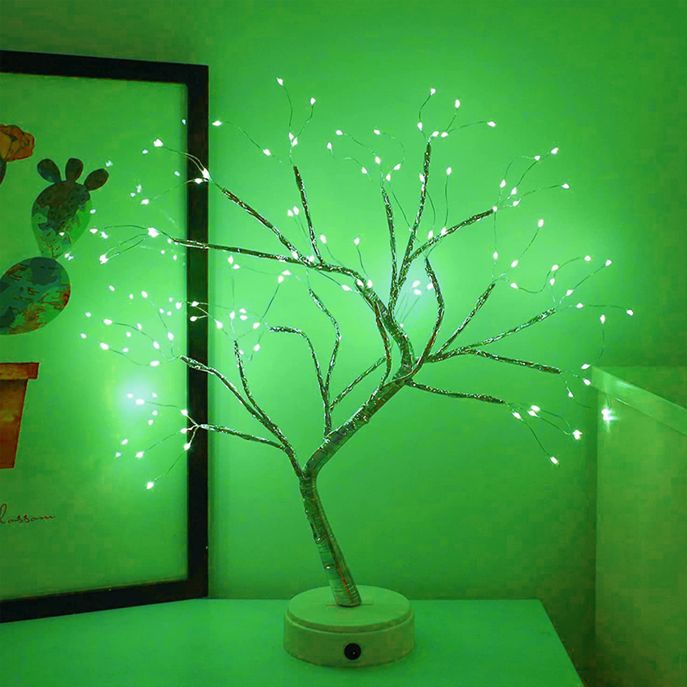 Ein dekorativer Baum mit kleinen weißen Lichtern steht auf einer grünen Oberfläche. Im Hintergrund befindet sich ein gerahmtes Bild mit einem Kaktus.