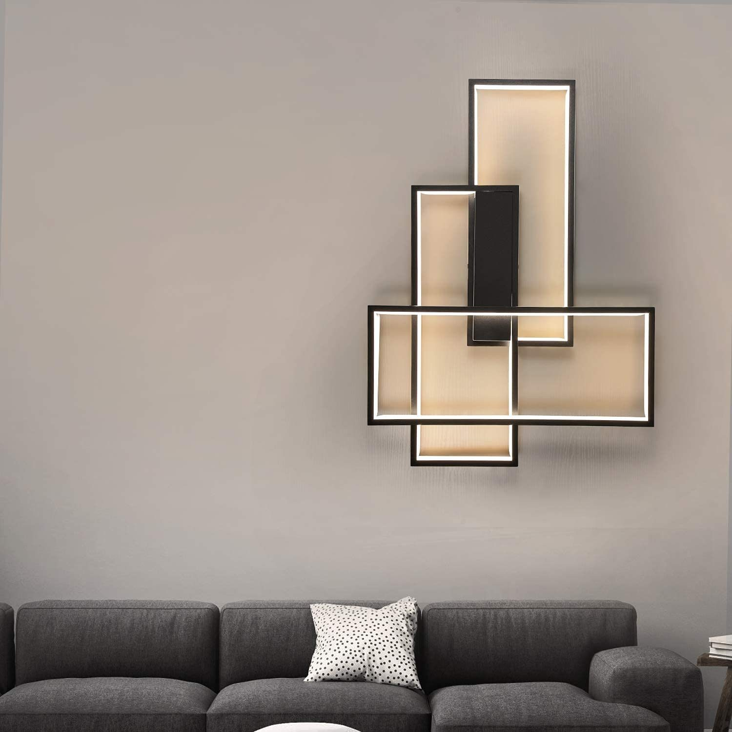 Lámpara de pared moderna con formas rectangulares, sobre un sofá gris con un cojín de lunares. Fondo de pared beige.