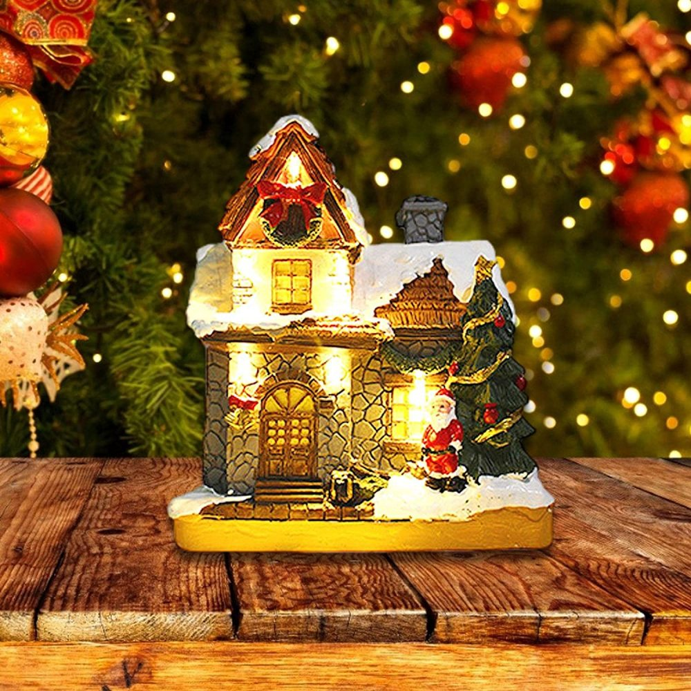 Eine Weihnachtsdorf-Szene. Ein Steinhaus mit Schnee, Santa und einem Baum. Auf einem Holztisch, mit Lichtern und Ornamenten im Hintergrund.