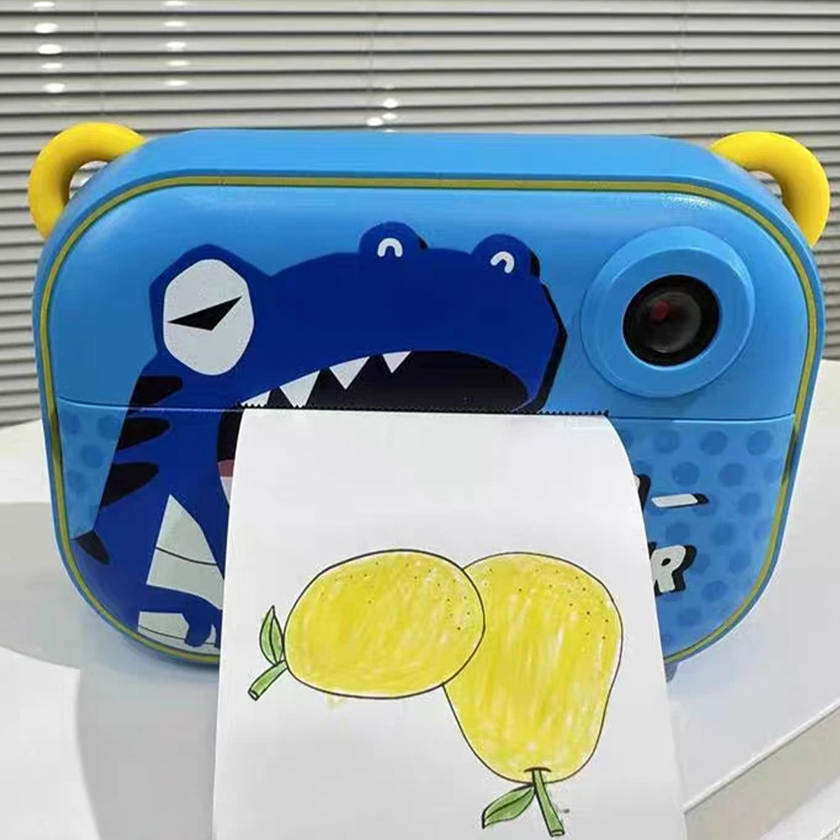 Una cámara de juguete azul con diseño de cocodrilo imprime un dibujo de dos limones amarillos en papel blanco.