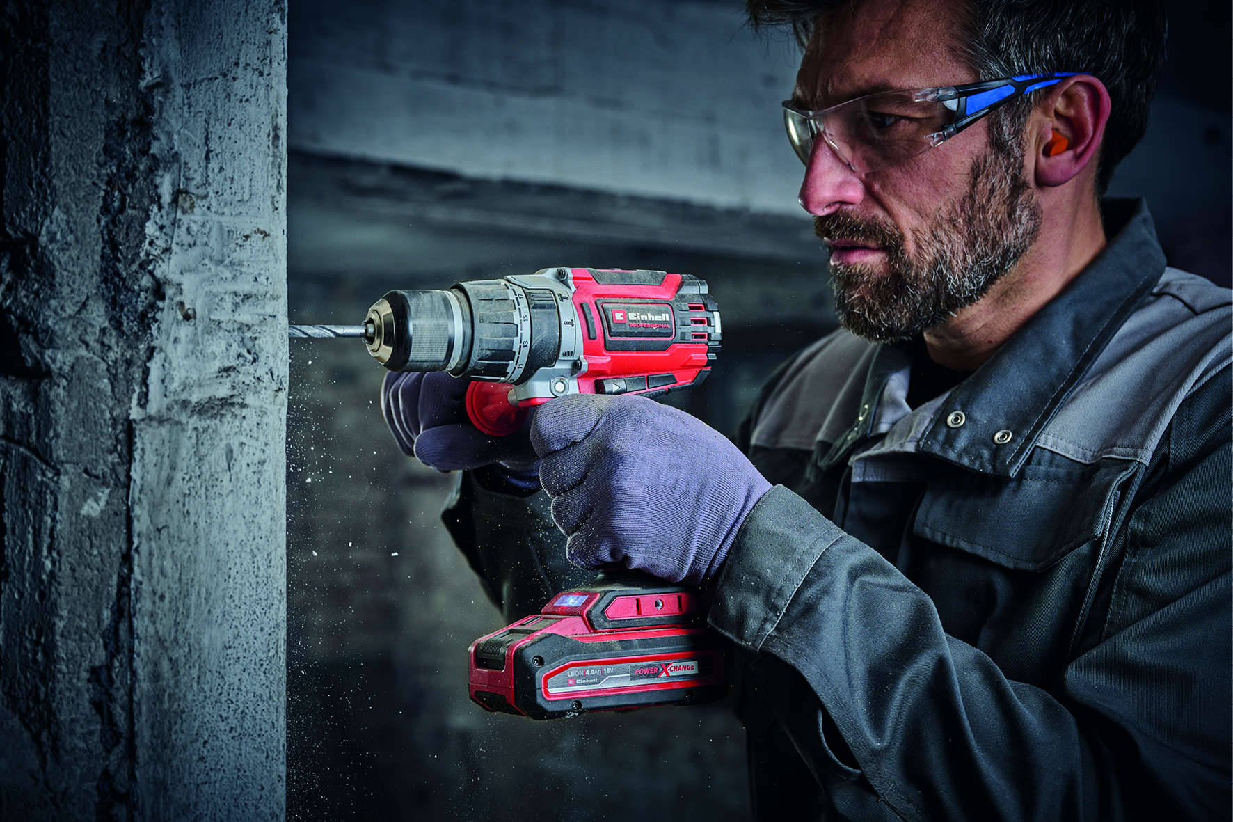 Ein Mann bohrt mit einem rot-schwarzen Elektrowerkzeug in Beton, trägt Handschuhe und eine Schutzbrille. Staub fliegt. Hintergrund ist eine Wand.