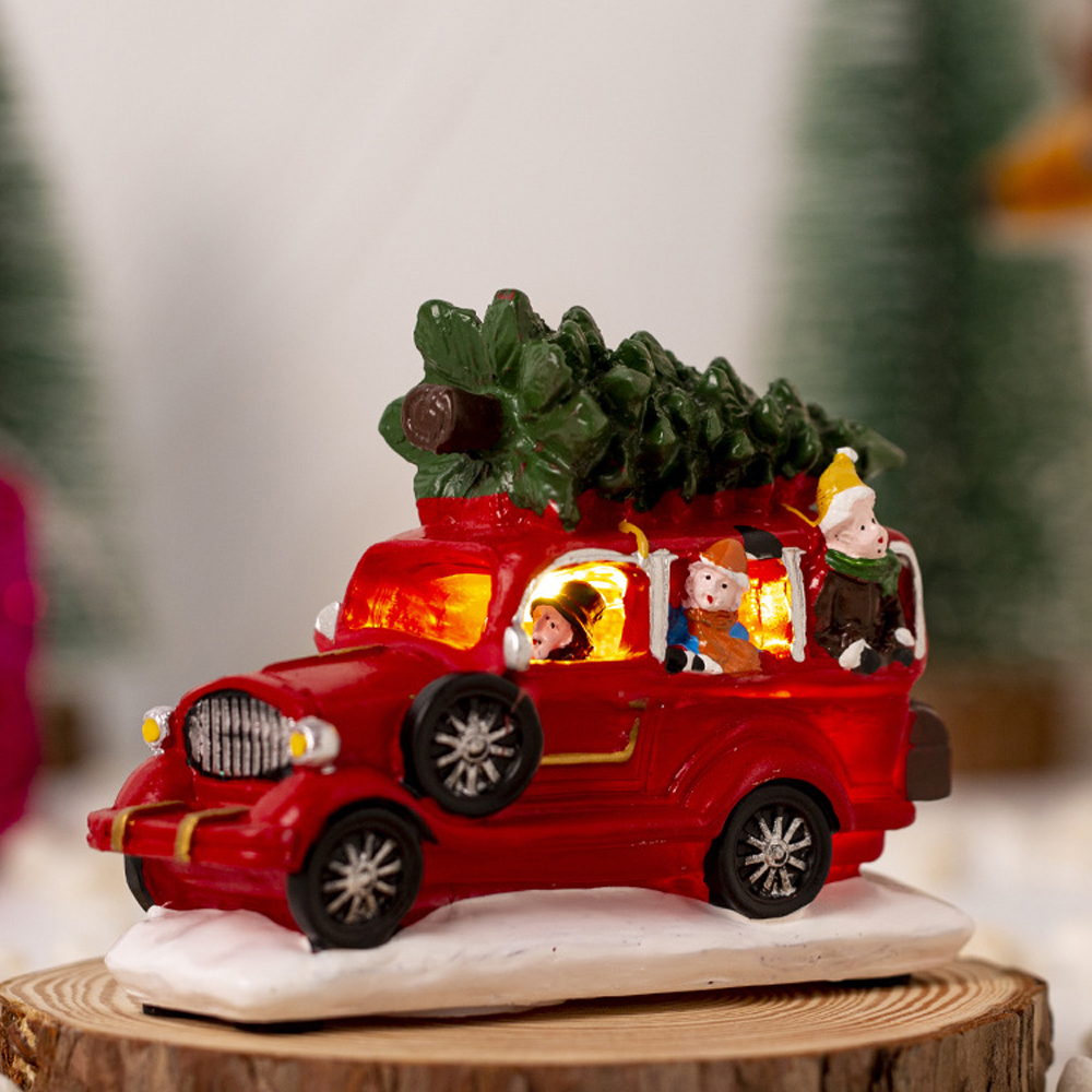 Ein rotes Spielzeugauto mit einem Weihnachtsbaum oben drauf. Im Inneren befinden sich Passagiere. Es steht auf einem Holzsockel mit Schnee.