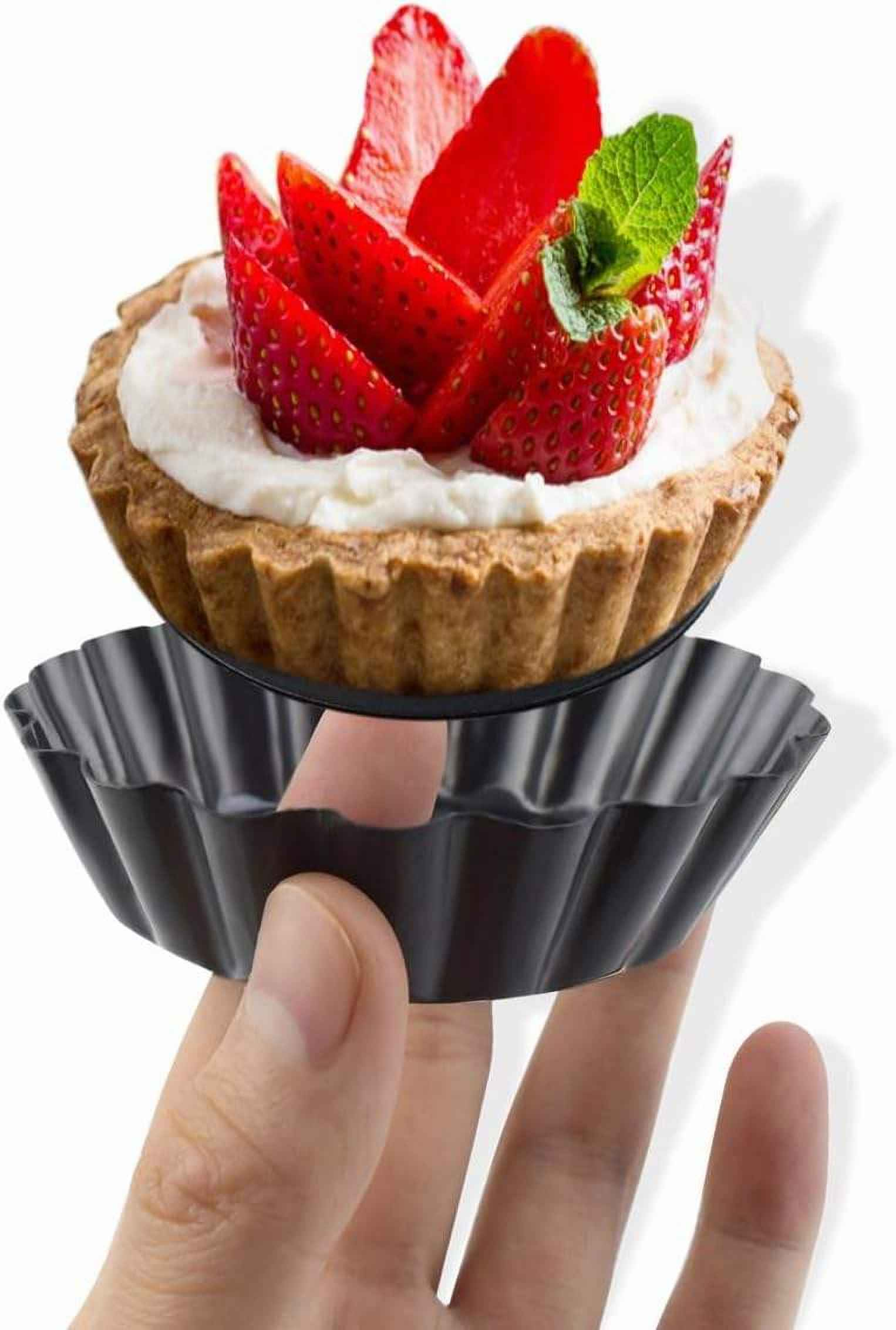 Eine Hand hält eine Tartelette-Pfanne mit einem Erdbeerkuchen darauf. Der Kuchen hat Erdbeeren und Sahne. Weißer Hintergrund.
