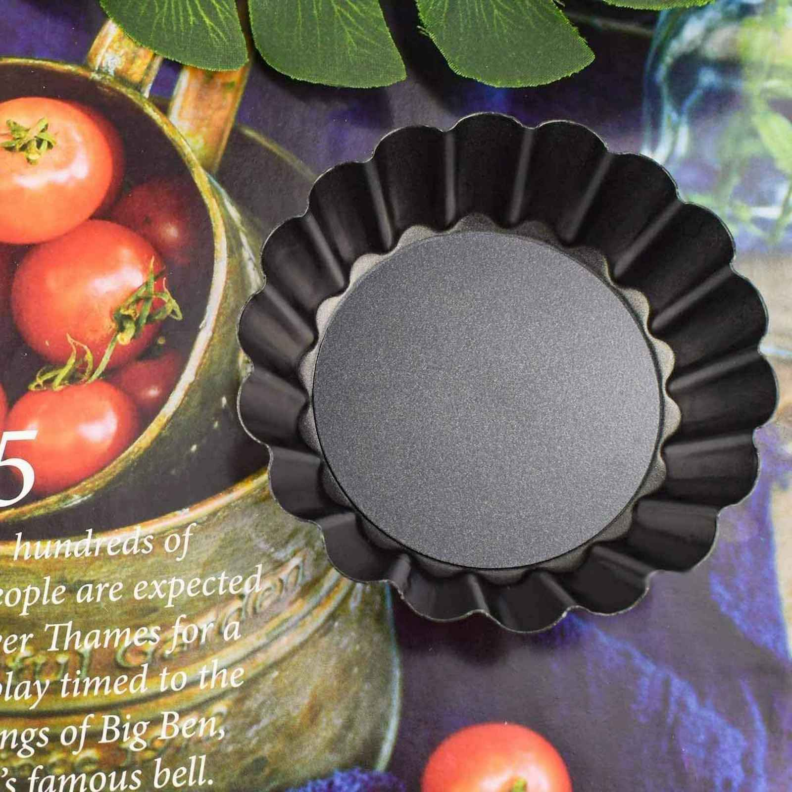Eine dunkle, geriffelte Tarteform liegt auf einer bunten Oberfläche, mit Tomaten in einer Schüssel und Text im Hintergrund.