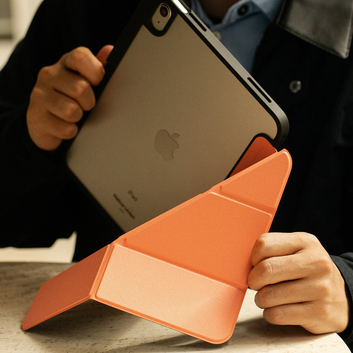Una persona tiene in mano un tablet con una cover arancione. Il tablet ha il logo Apple. Lo sfondo è scuro, con uno scorcio di camicia azzurra.