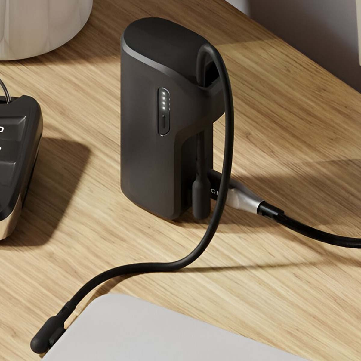 Une batterie externe noire avec un câble connecté, posée sur une surface en bois. Un clavier est à gauche, et un pot blanc en haut à gauche.