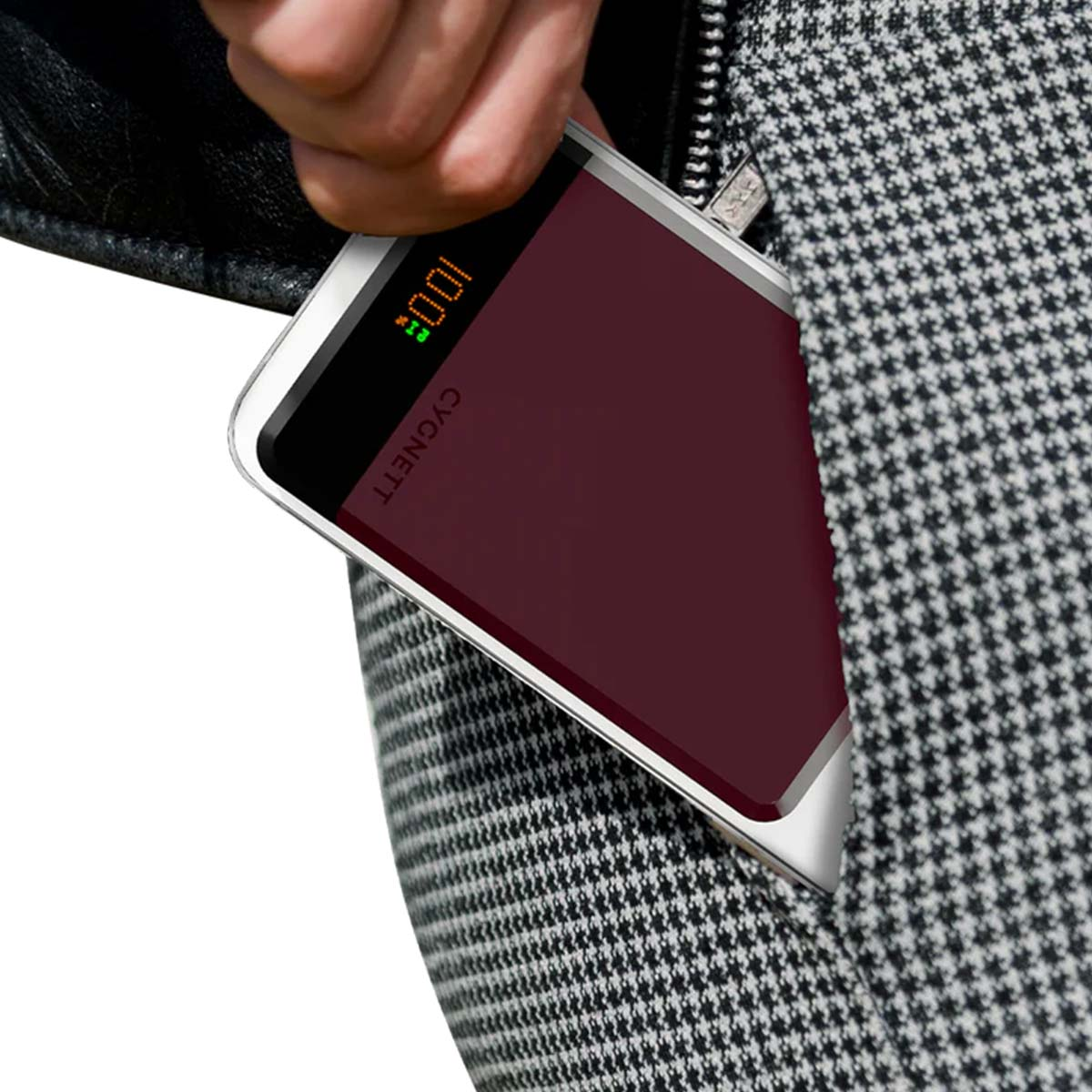 Una mano tiene in mano un power bank Cygnnet. Il dispositivo ha una superficie bordeaux, che mostra il 100% di carica. Lo sfondo è una giacca a fantasia.