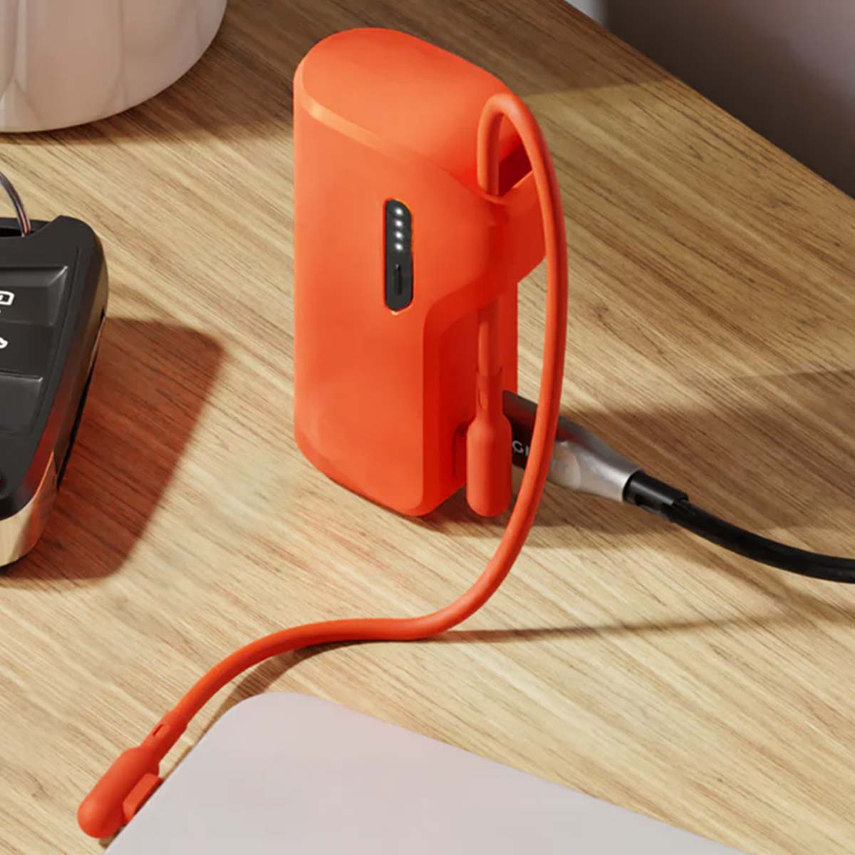 Eine orangefarbene Powerbank mit angeschlossenem Kabel neben einem Autoschlüsselanhänger auf einer Holzoberfläche. Die Powerbank hat eine Ladeanzeige.