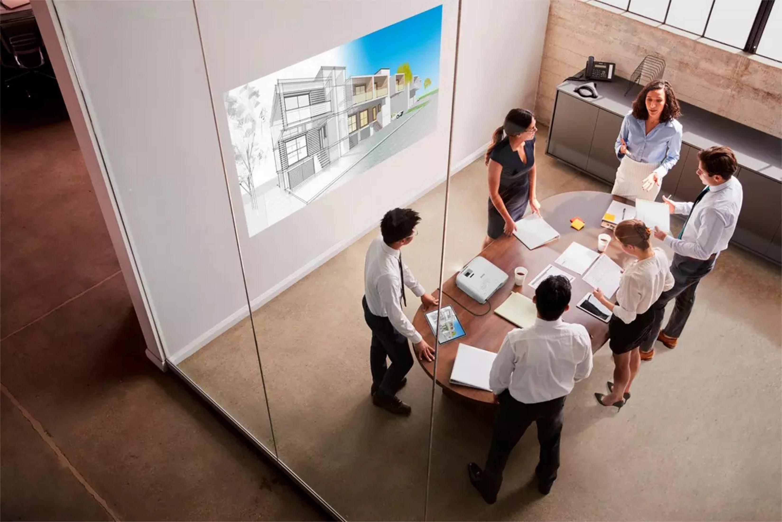 Spotkanie biznesowe w nowoczesnym biurze. Projekt budowlany wyświetlany na ścianie. Ludzie wokół okrągłego stołu.