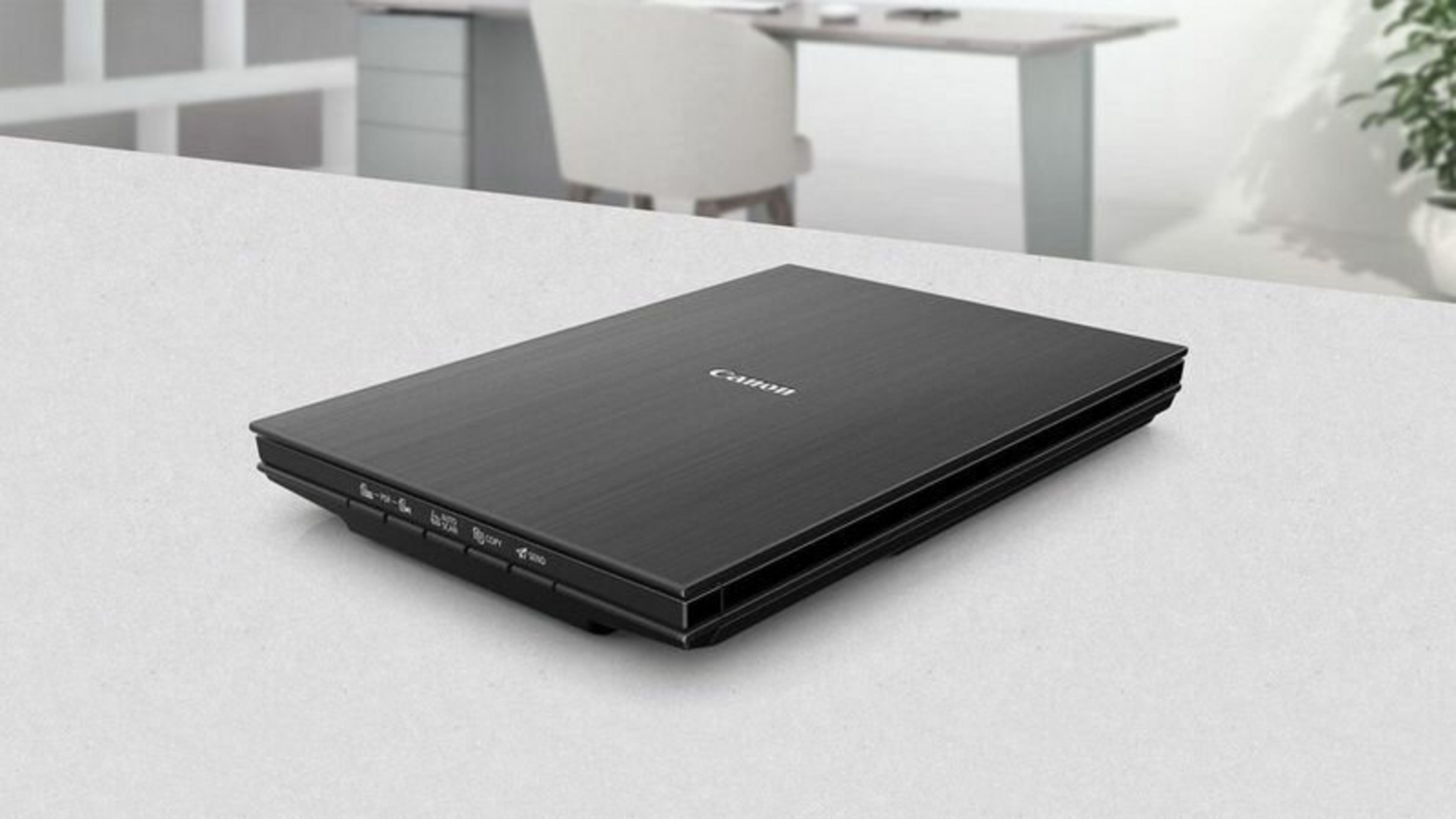 Skaner Canon leży na białej powierzchni. W tle biurko, krzesło i roślina. Skaner jest czarny.