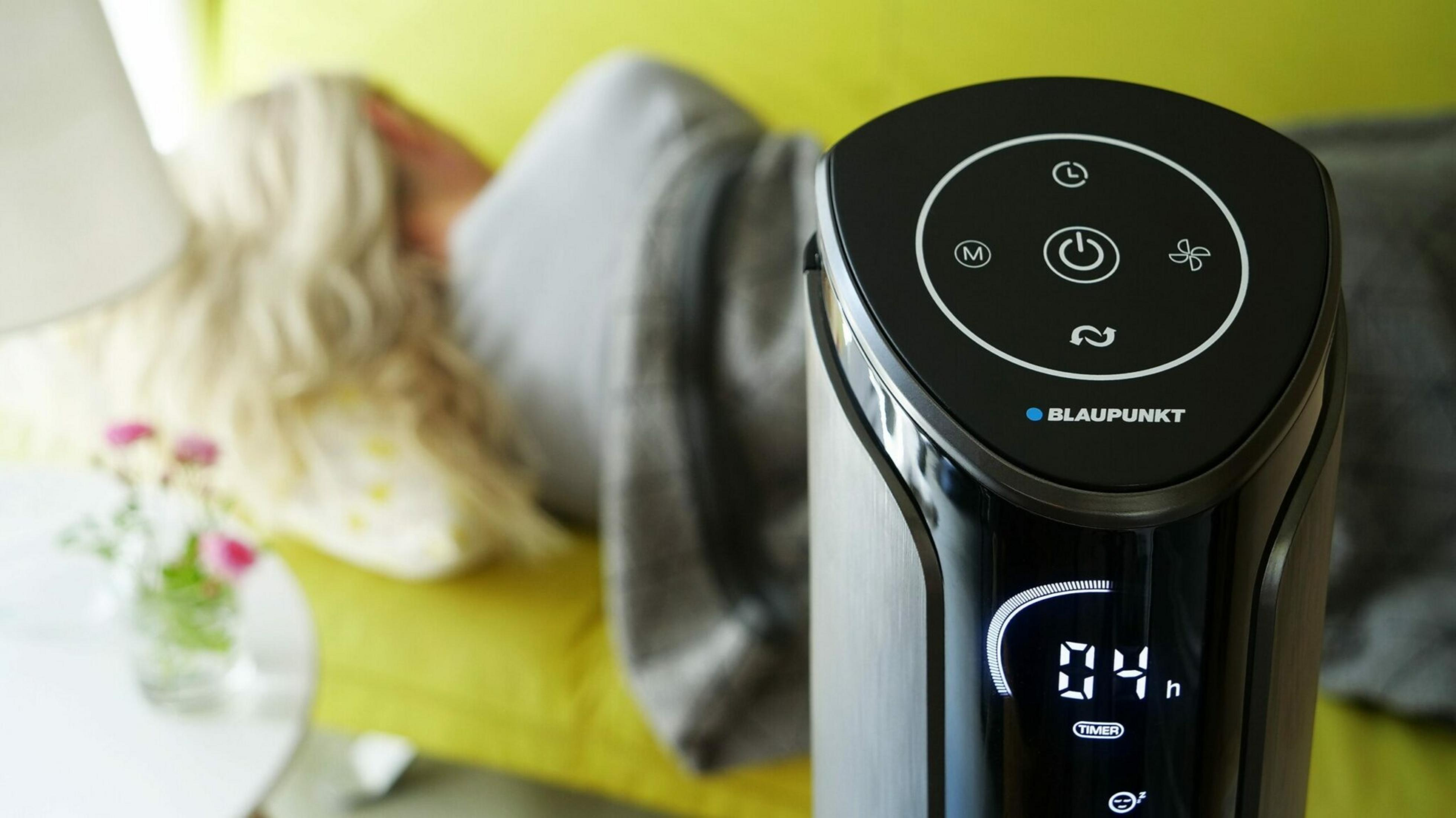 Un purificador de aire Blaupunkt está encendido con temporizador. Una persona está acostada en un sofá amarillo en el fondo borroso.