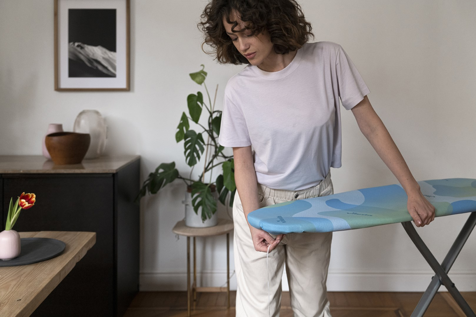 Kobieta trzyma deskę do prasowania. W tle roślina i obraz w ramie.