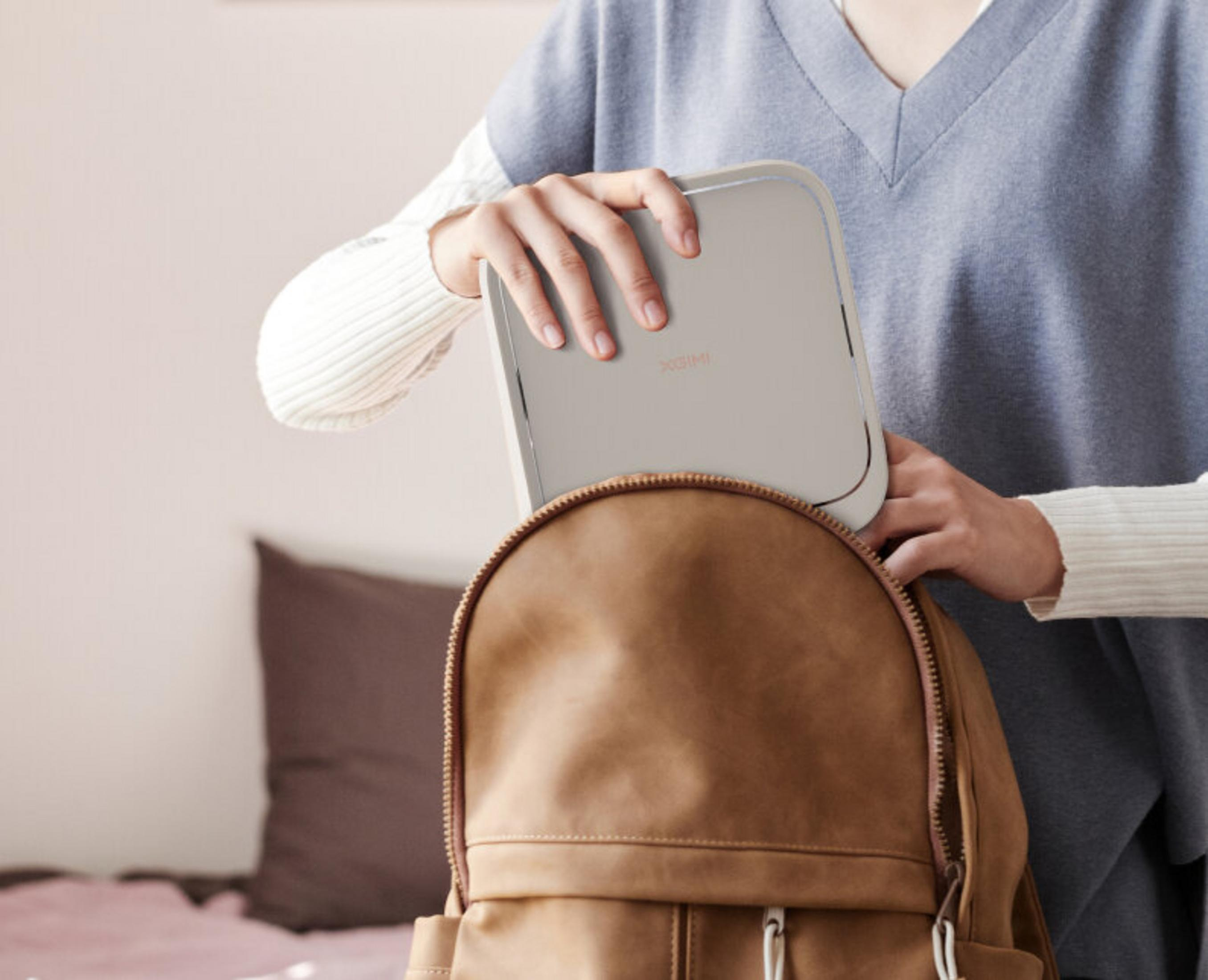 Osoba umieszcza szare urządzenie w brązowym plecaku. Osoba ma na sobie niebieski sweter i białe rękawy.