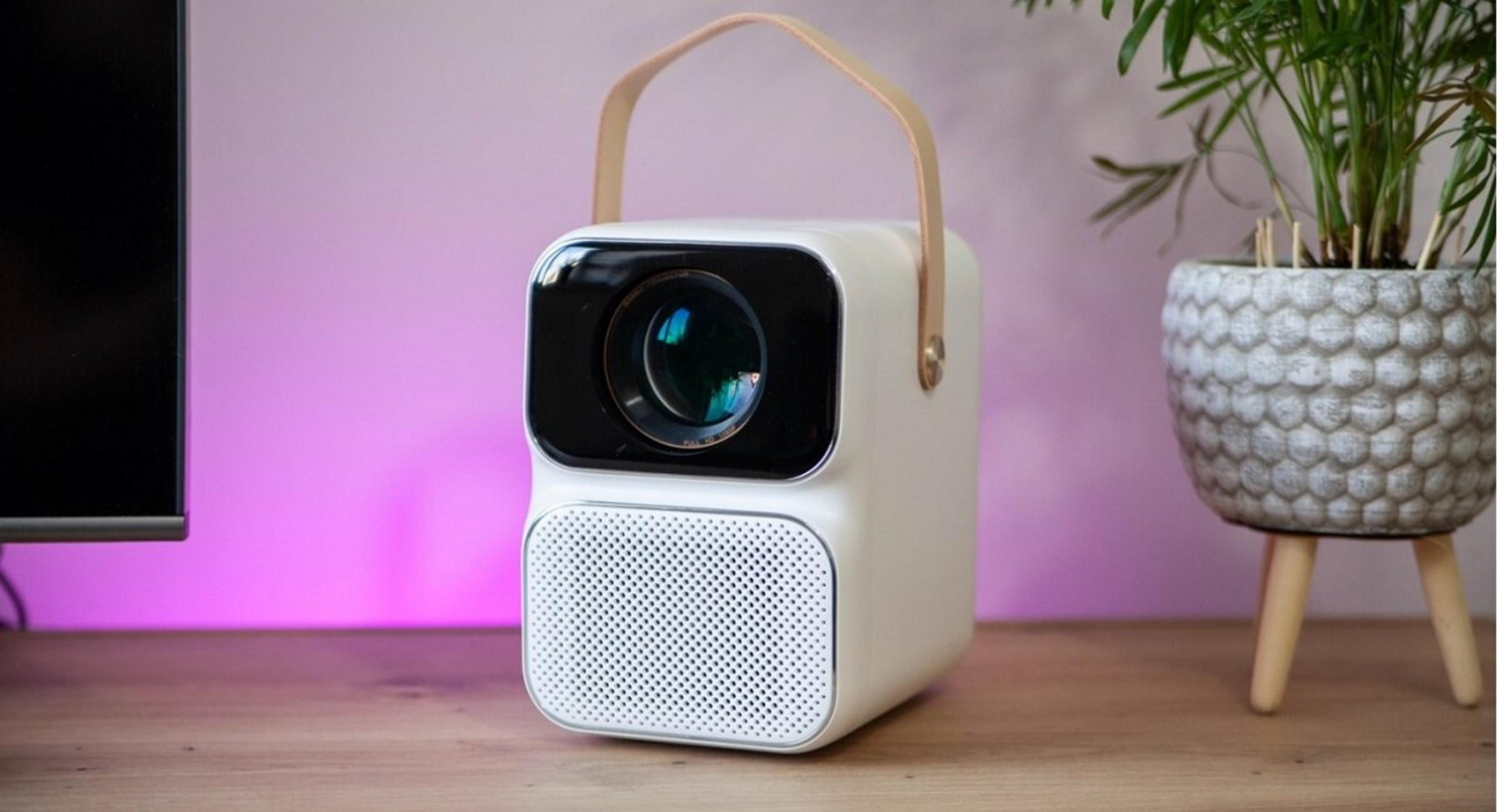 Un proyector blanco con asa sobre una superficie de madera. A la derecha, una planta en maceta gris.