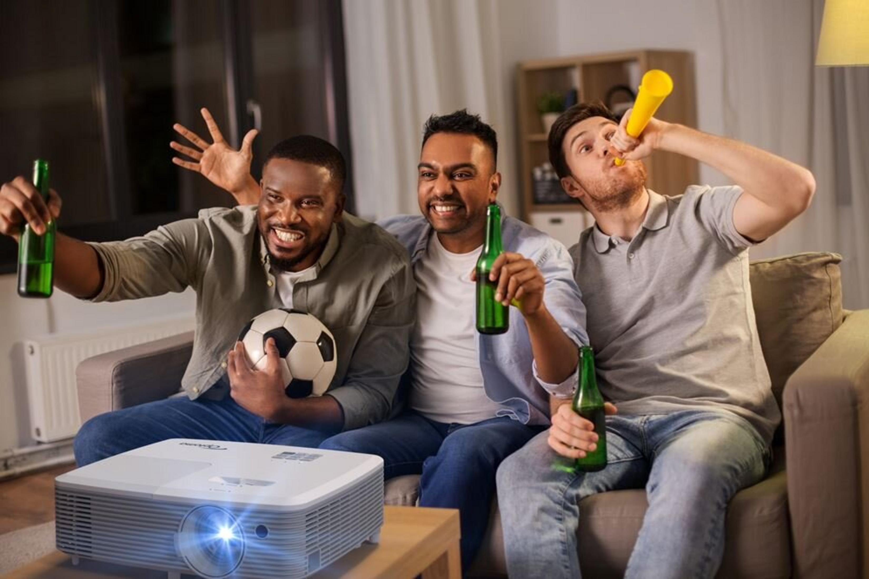 Tres hombres ven un partido, sostienen cerveza y animan. Uno toca una bocina, otro sostiene un balón.