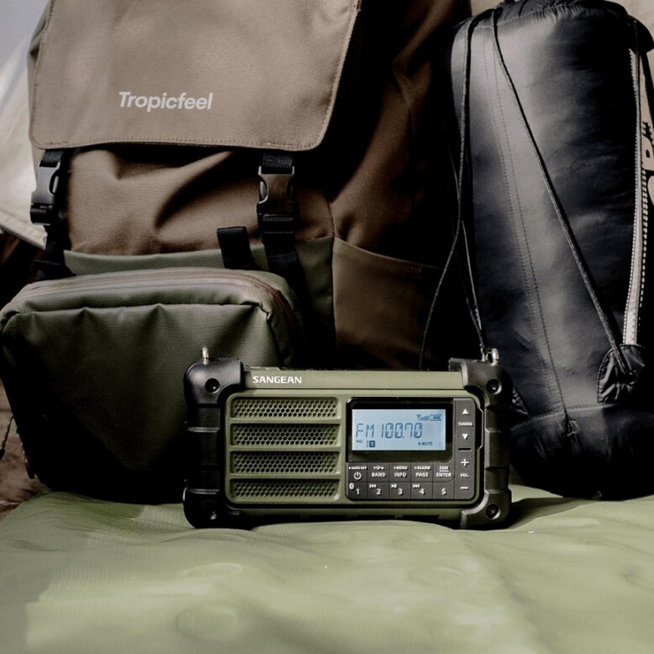 Una radio verde oliva mostra FM 100.70. Uno zaino, una borsa e un sacco a pelo sono dietro, su una superficie verde.