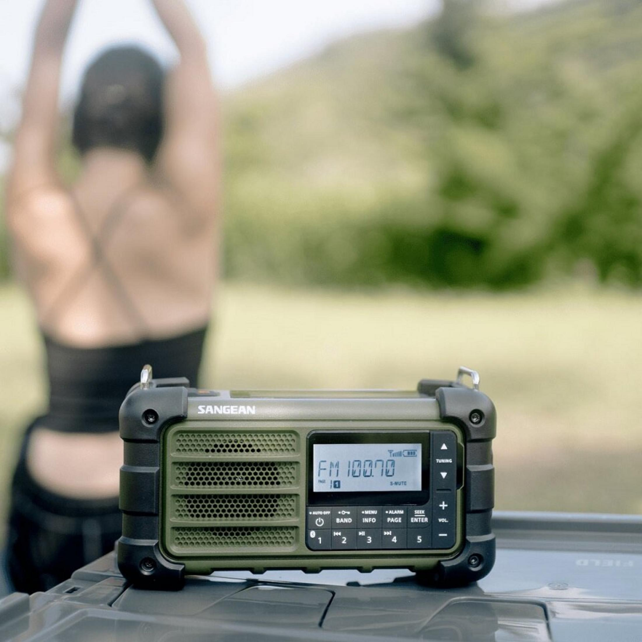 Una persona si allunga vicino a una radio. La radio mostra FM 100.70. Erba verde e alberi sullo sfondo.