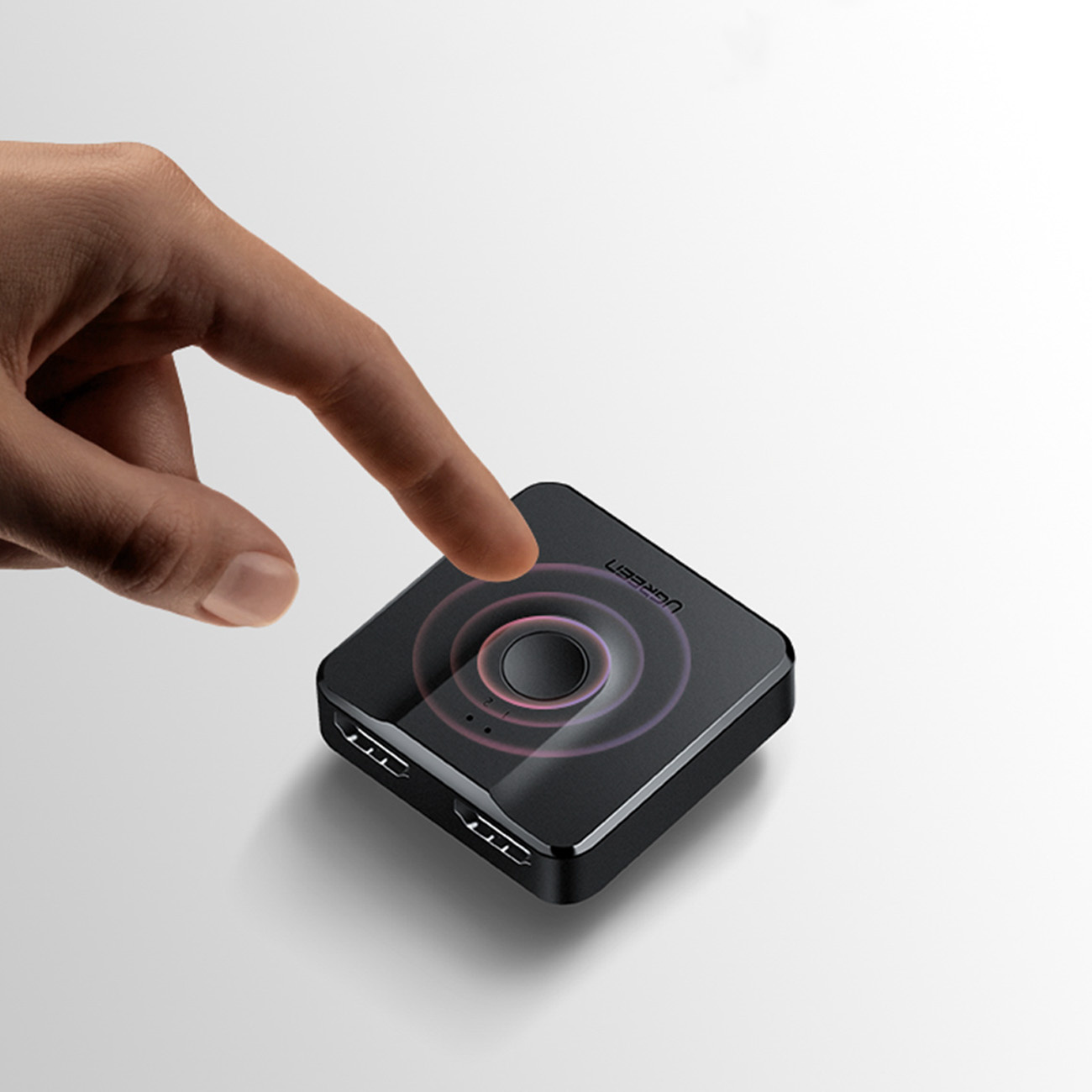 Dłoń dotyka czarnego kwadratowego urządzenia. Urządzenie ma koncentryczne okręgi i napis UGREEN. Białe tło.