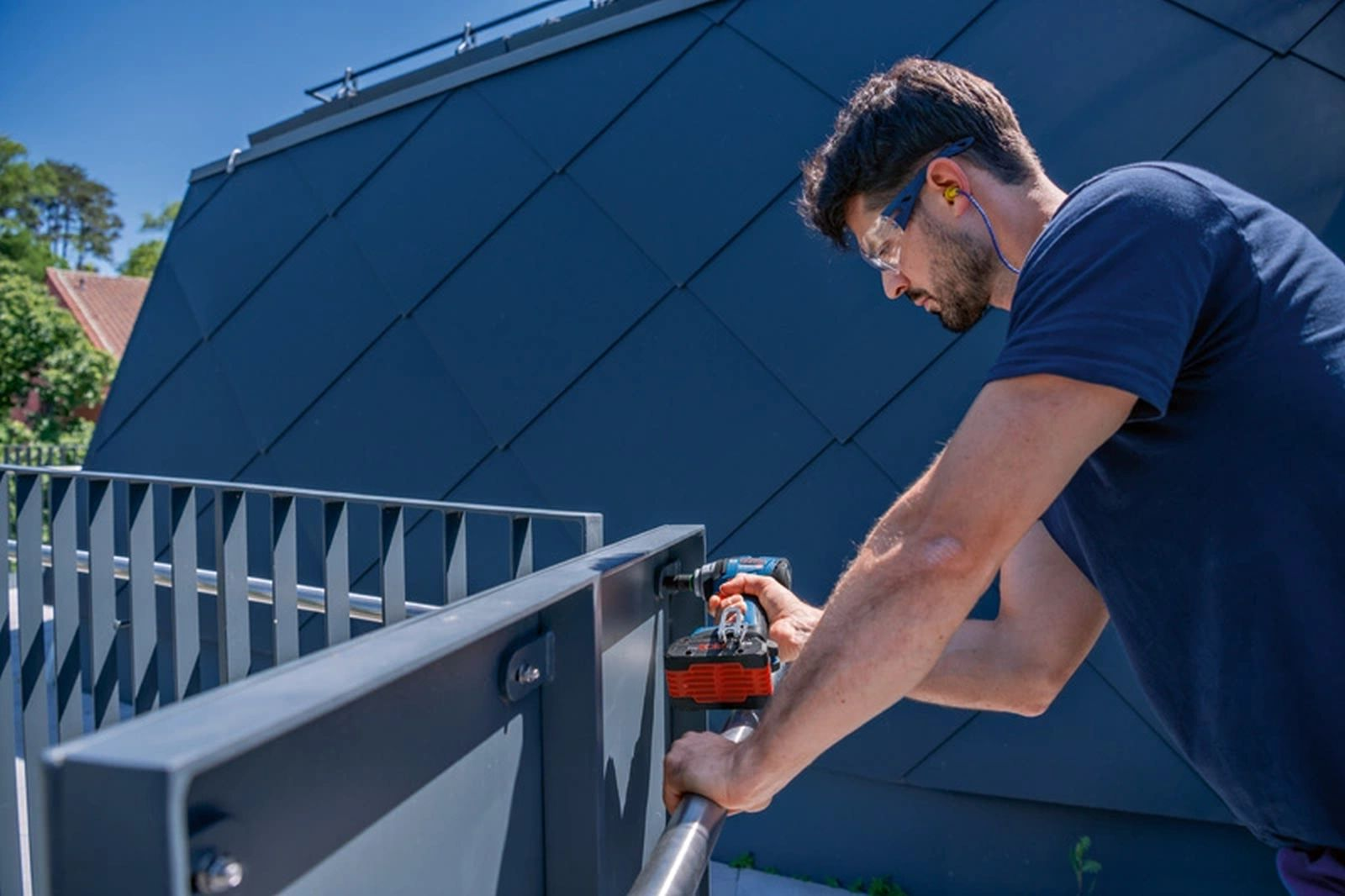 Mężczyzna w okularach ochronnych wierci w metalowej balustradzie. Tło: niebieski dach i słoneczne niebo.
