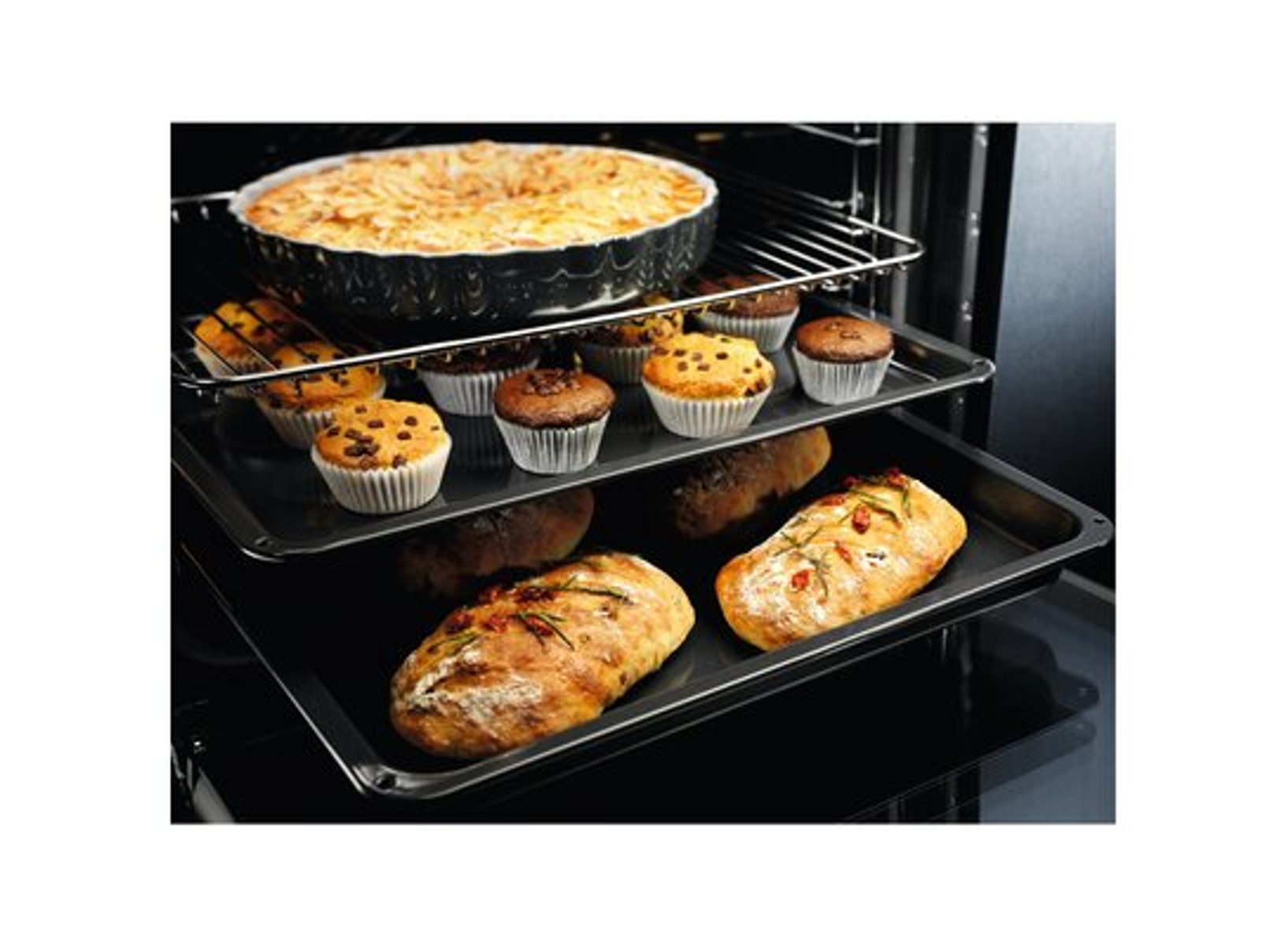 Piekarnik z wypiekami: ciasto, babeczki i chleb. Ciasto jest na górnym ruszcie. Babeczki i chleb są na dolnych rusztach.