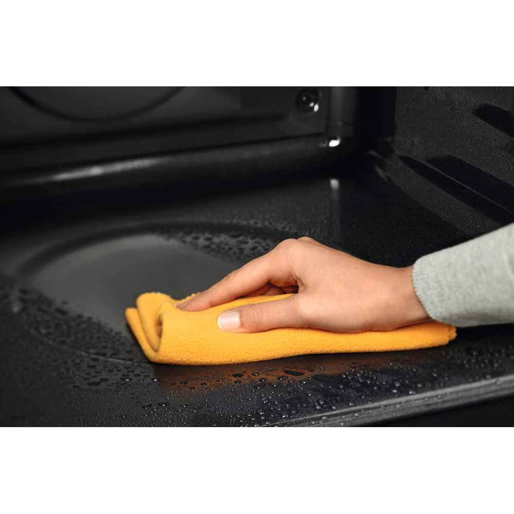 Ręka z żółtą szmatką wyciera wnętrze czarnego piekarnika. Krople wody są widoczne na powierzchni.