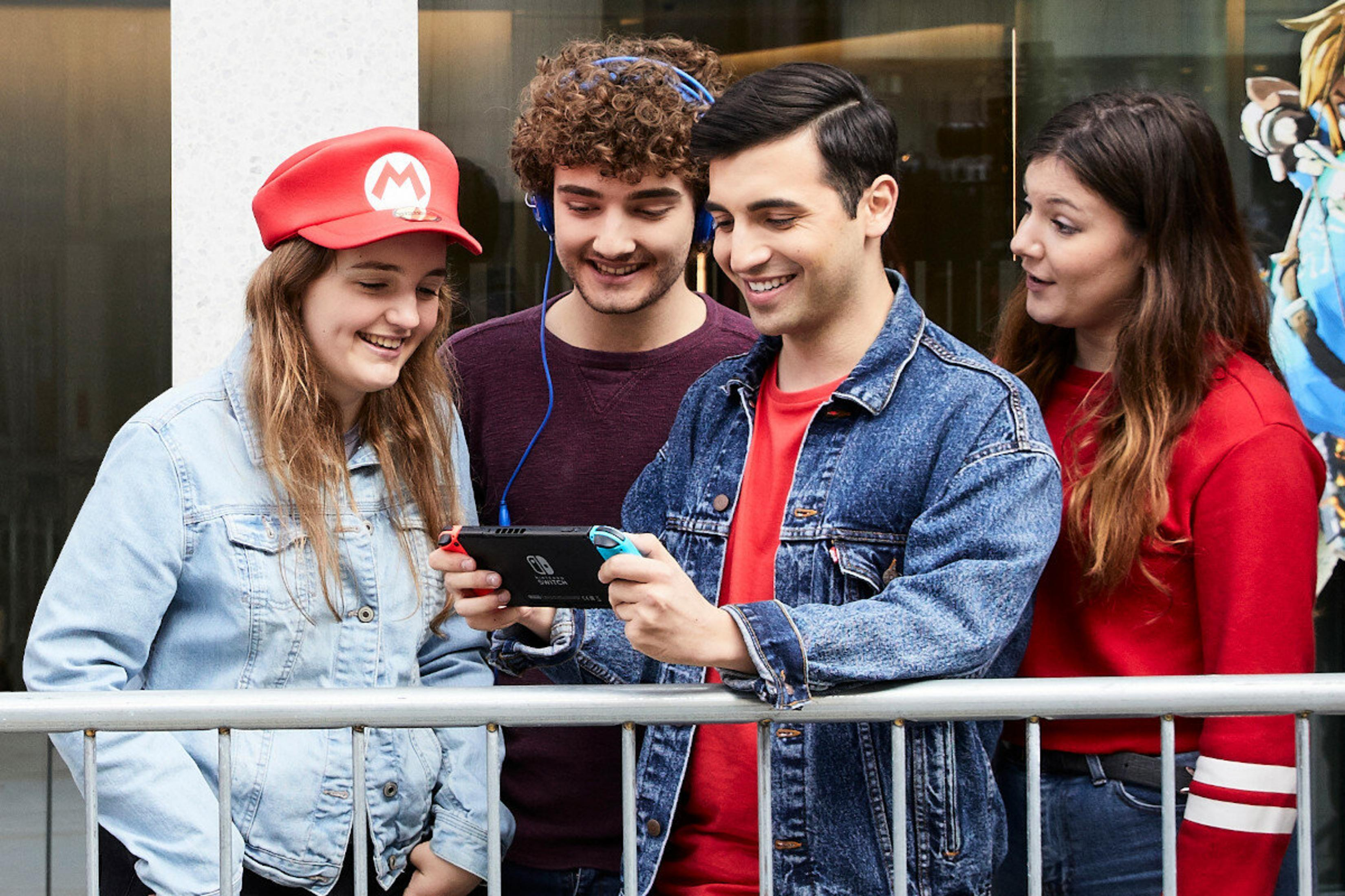 Vier junge Leute mit einer Nintendo Switch. Einer trägt einen Mario-Hut. Sie lächeln und schauen auf den Bildschirm.