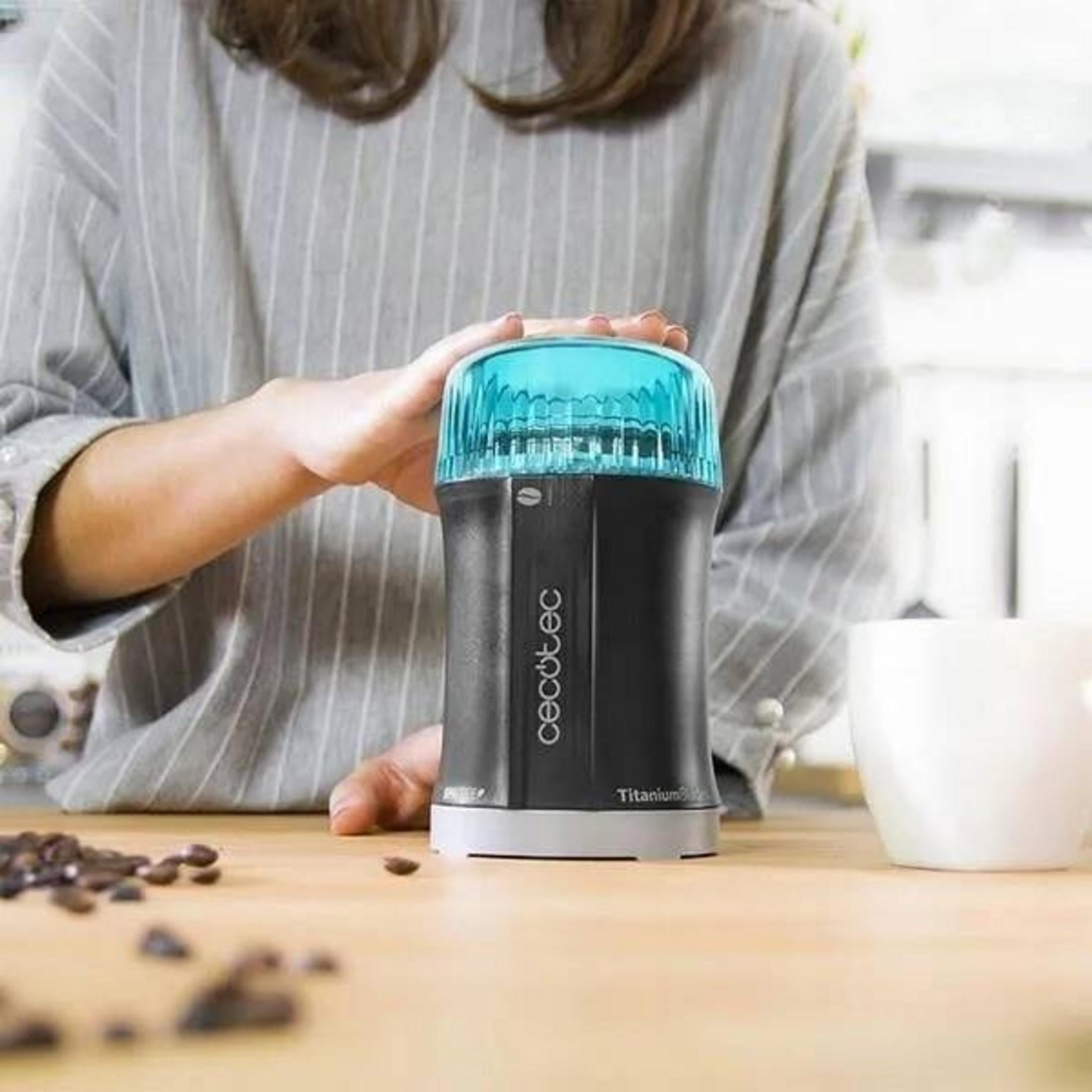 Una persona tiene in mano un macinacaffè nero con la parte superiore blu. Chicchi di caffè e una tazza bianca sulla superficie di legno.