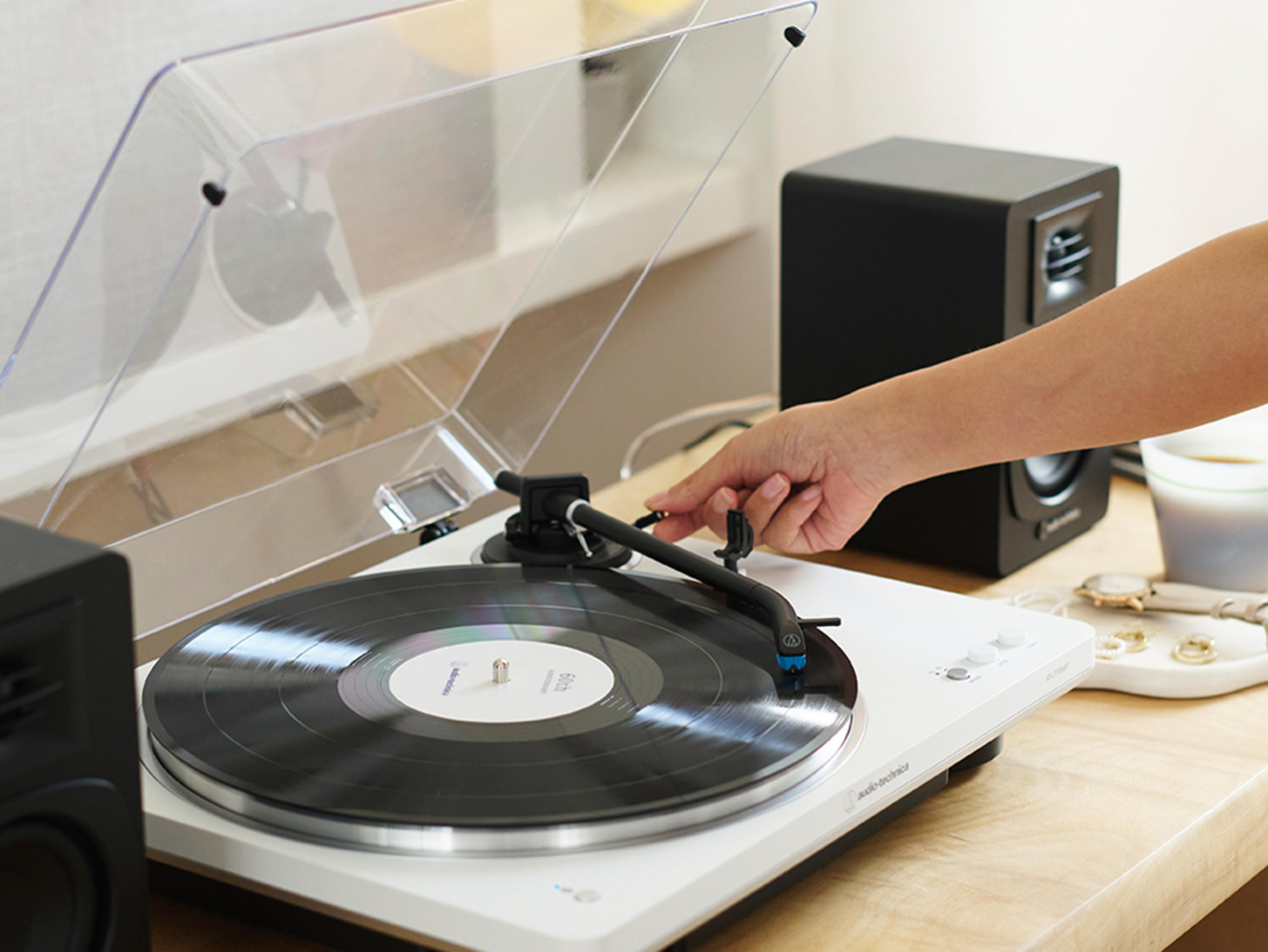 Ręka opuszcza ramię gramofonu. W pobliżu znajduje się czarny głośnik i akcesoria.