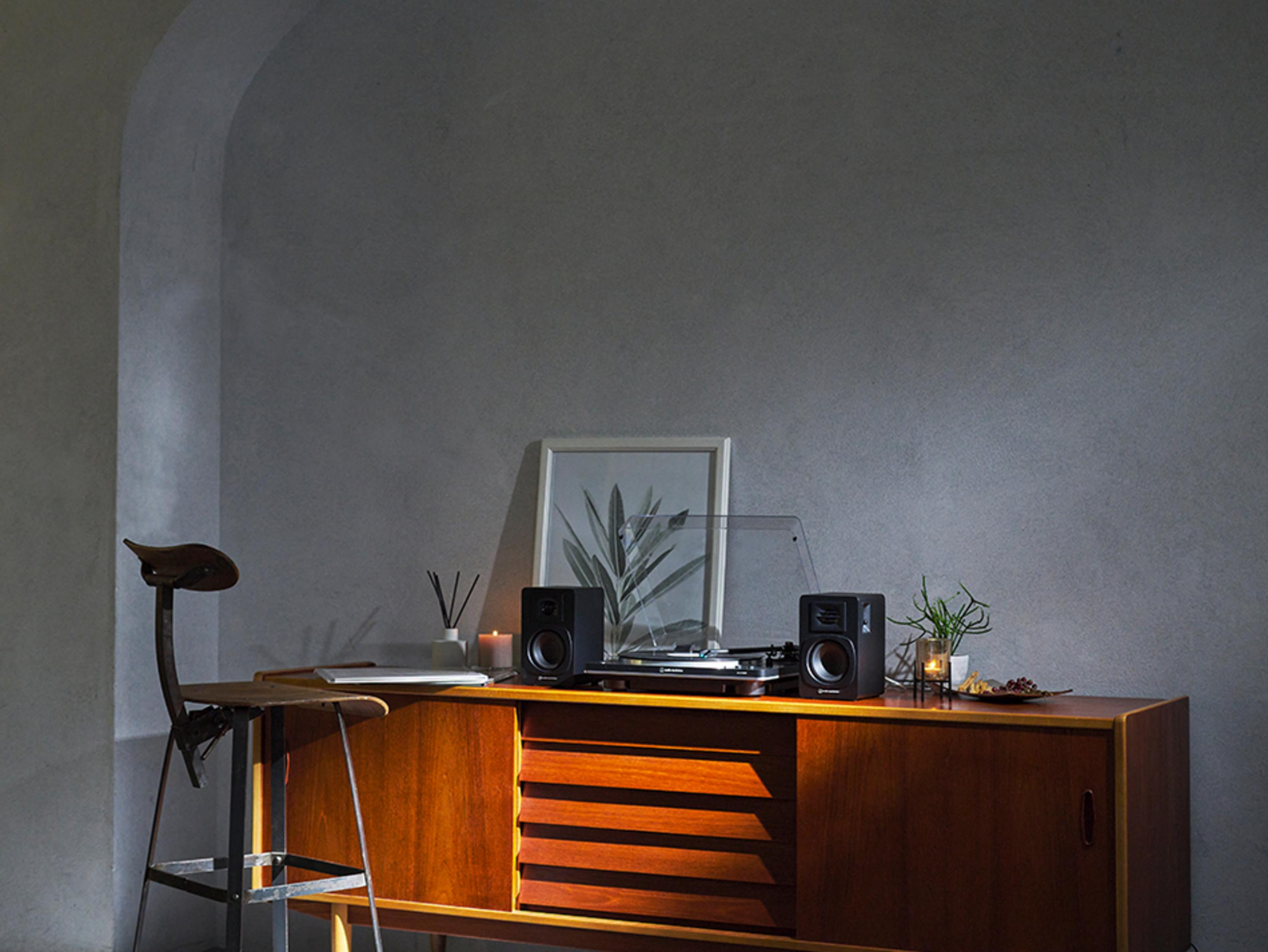 Pokój z drewnianą szafką, głośnikami, gramofonem, świecami i oprawionym obrazem, krzesło po lewej.